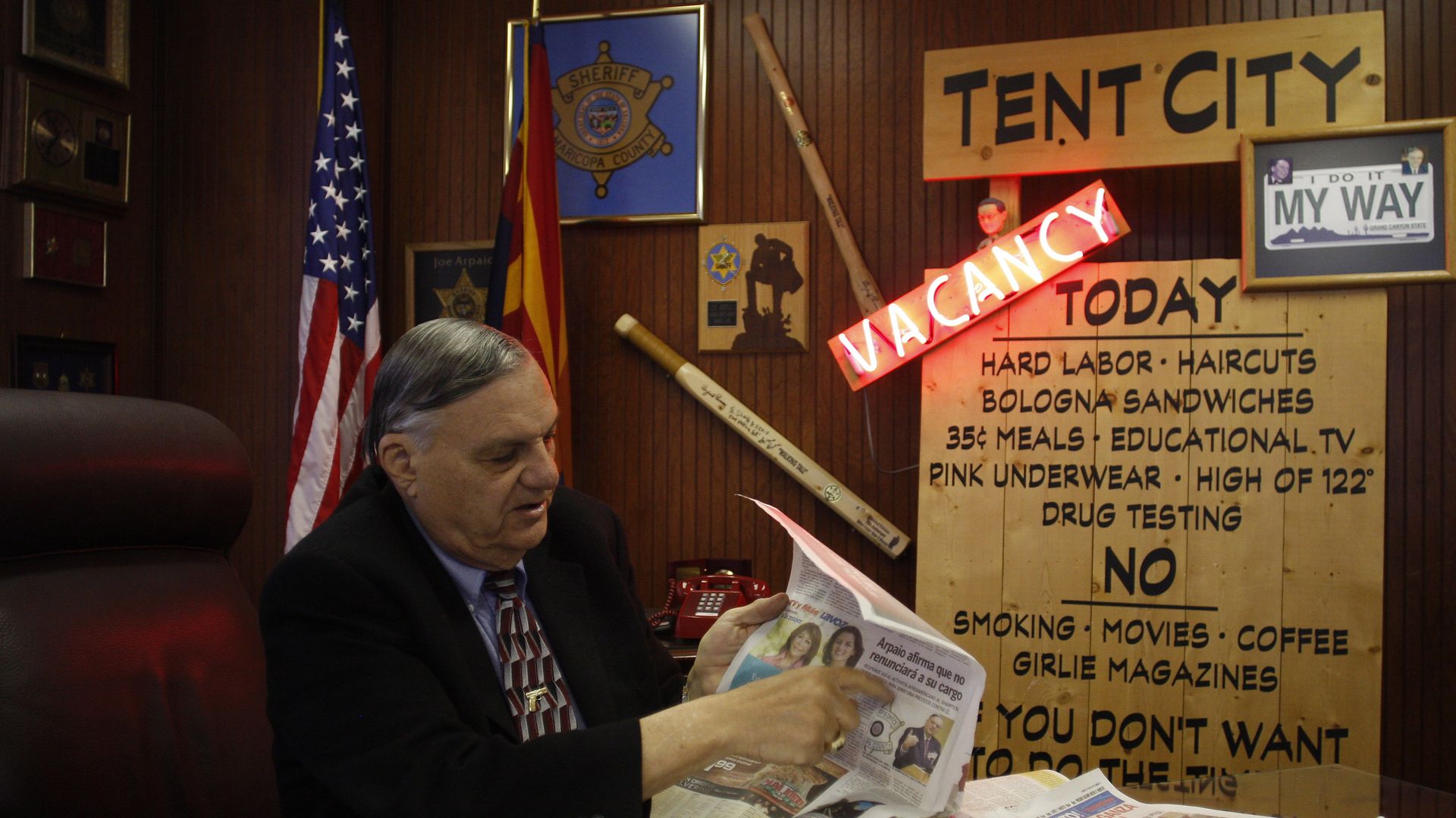 Man in a suit sitting at desk with newspapers, behind him flags and a sheriff sign. Large wooden board labeled "Tent City" listing rules and services, with a red "Vacancy" neon sign.
