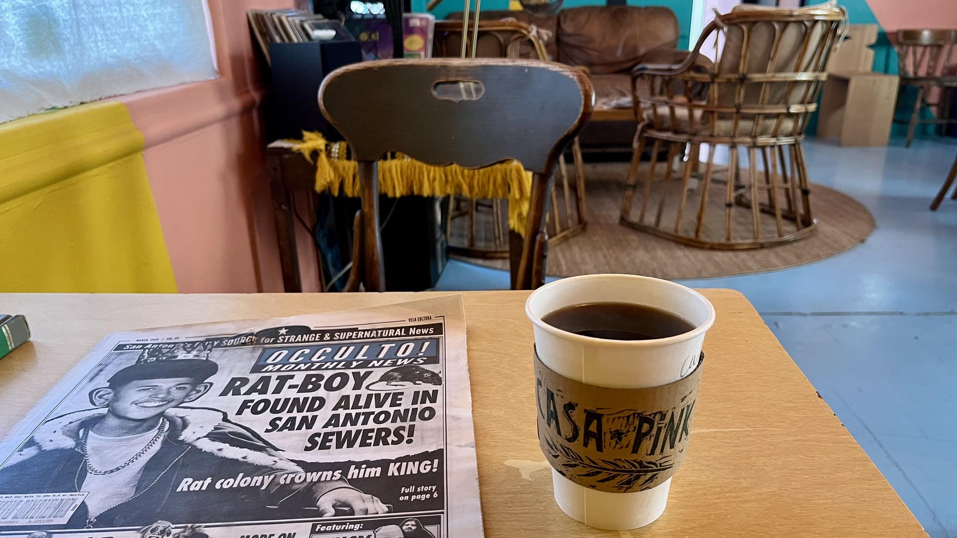 A wooden table in the colorful cafe at Casa Pink Studio Coffee with a copy of Occulto!", a mock tabloid of San Antonio horror subculture, on it. The headline reads "Rat-Boy found alive in San Antonio sewers" and sits next to a takeaway coffee cup.