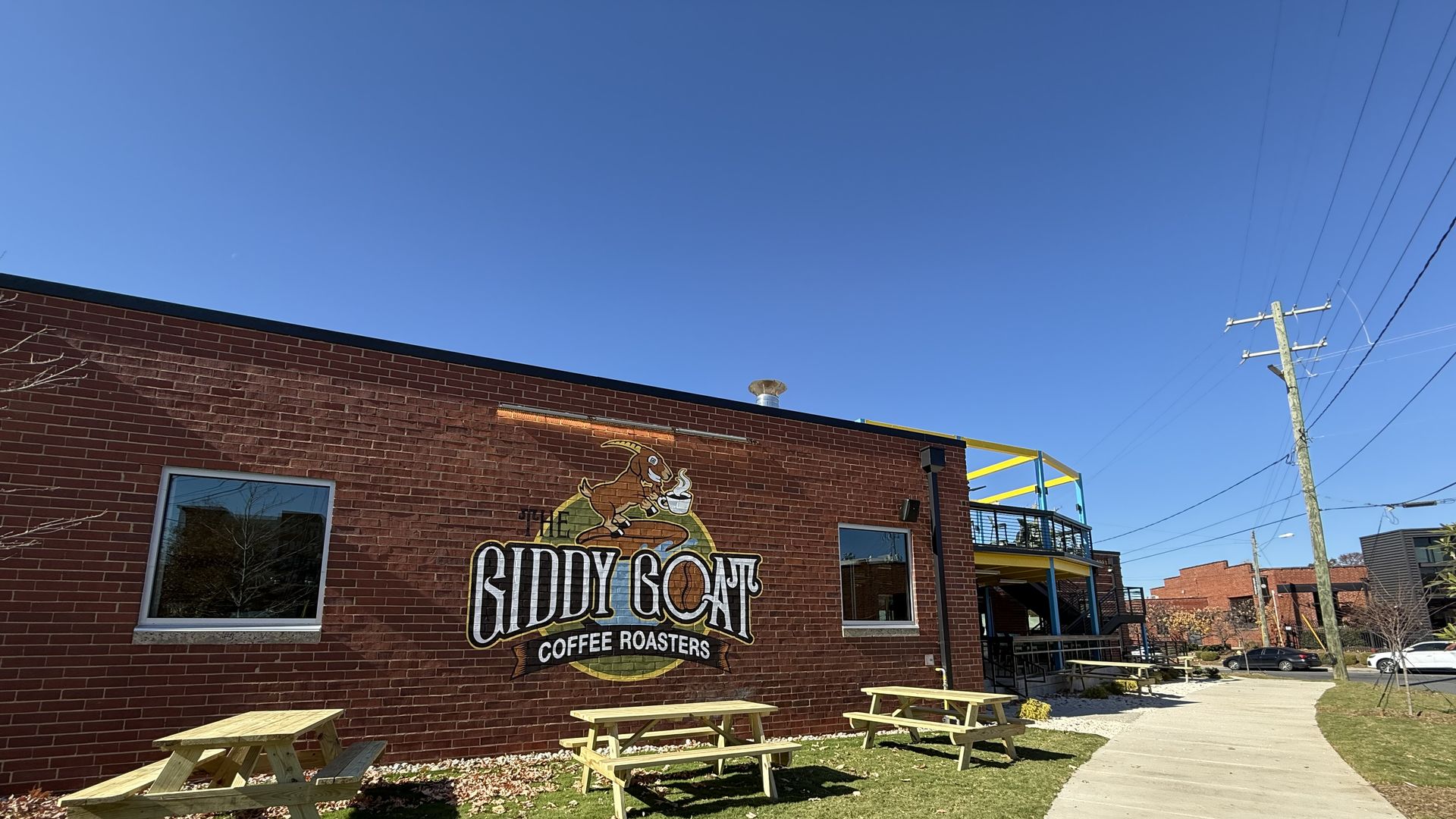 Red brick building with "The Giddy Goat Coffee Roasters" painted logo, outdoor wooden picnic tables on green grass, clear blue sky, and a sidewalk with a utility pole.