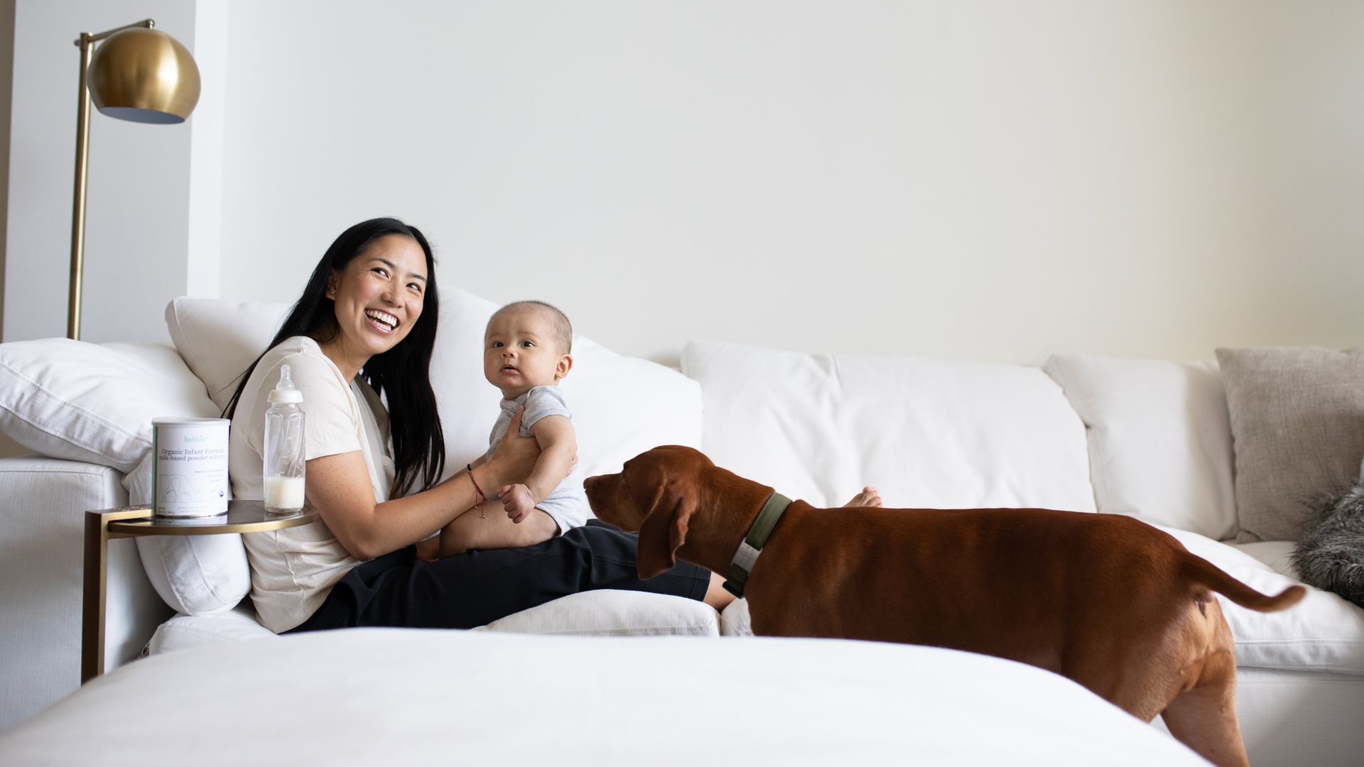 A smiling woman with long black hair holds an infant while reclining on a white sofa. Next to her on a tray table sits a package of infant formula and a baby bottle. A brown dog investigates..