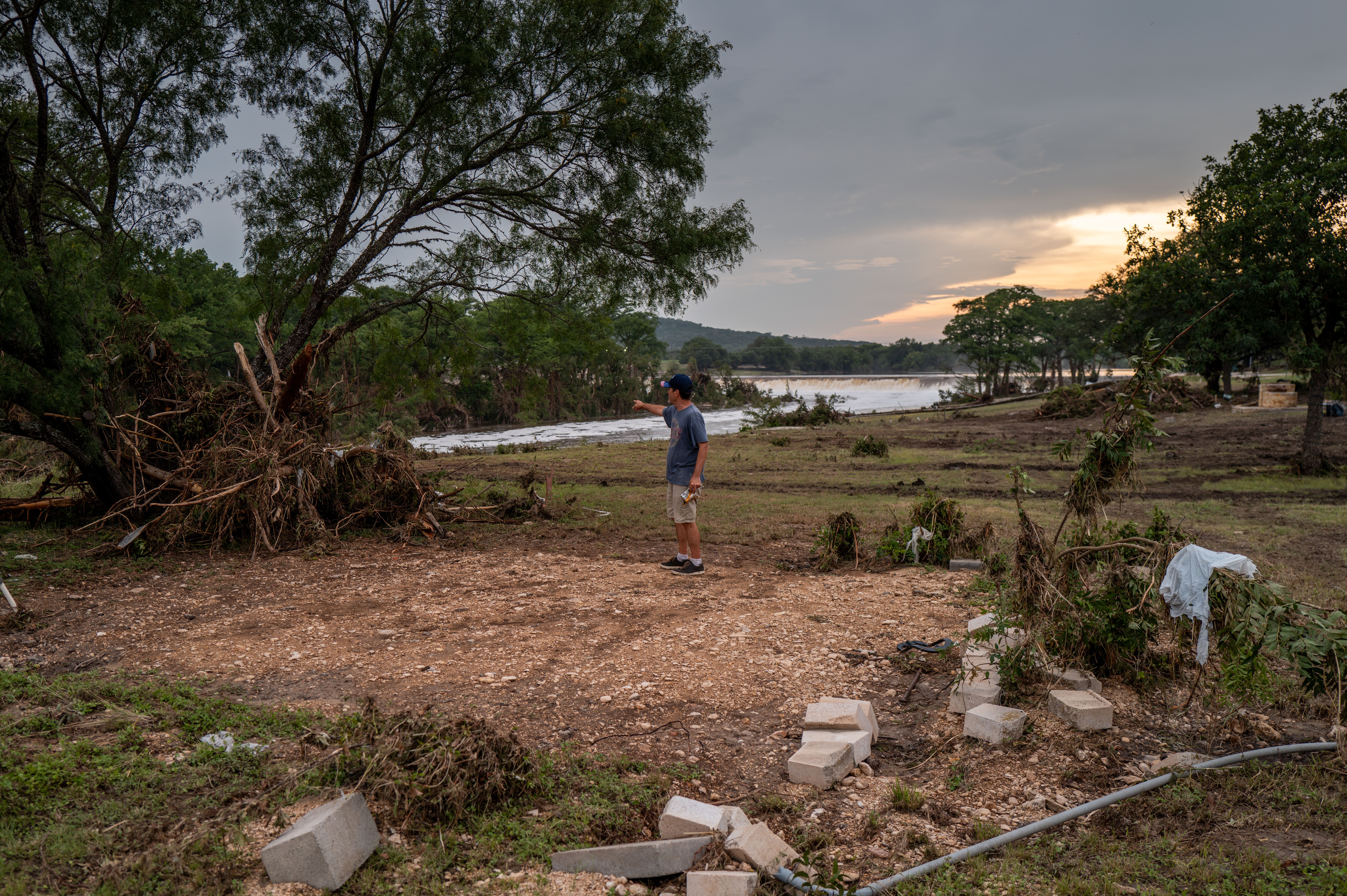A man is seen pointing and standing in a rectangle of debris-filled land surrounded by grass and an uprooted tree, with the Guadalupe River in the background. The rectangle of land where he stands is where his living room once was. His home was swept away during the storm.