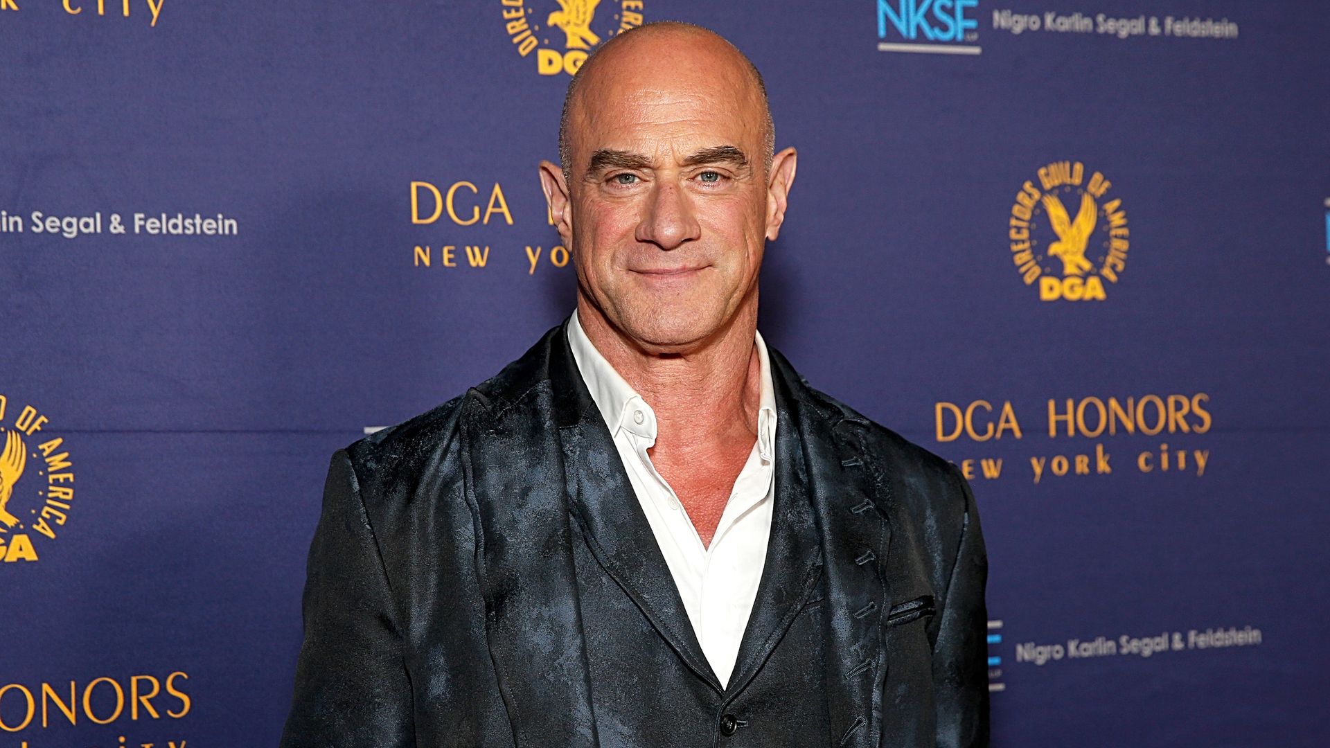 A bald man wearing a dark, shiny suit jacket and white shirt poses in front of a blue backdrop with gold DGA logos and event text.