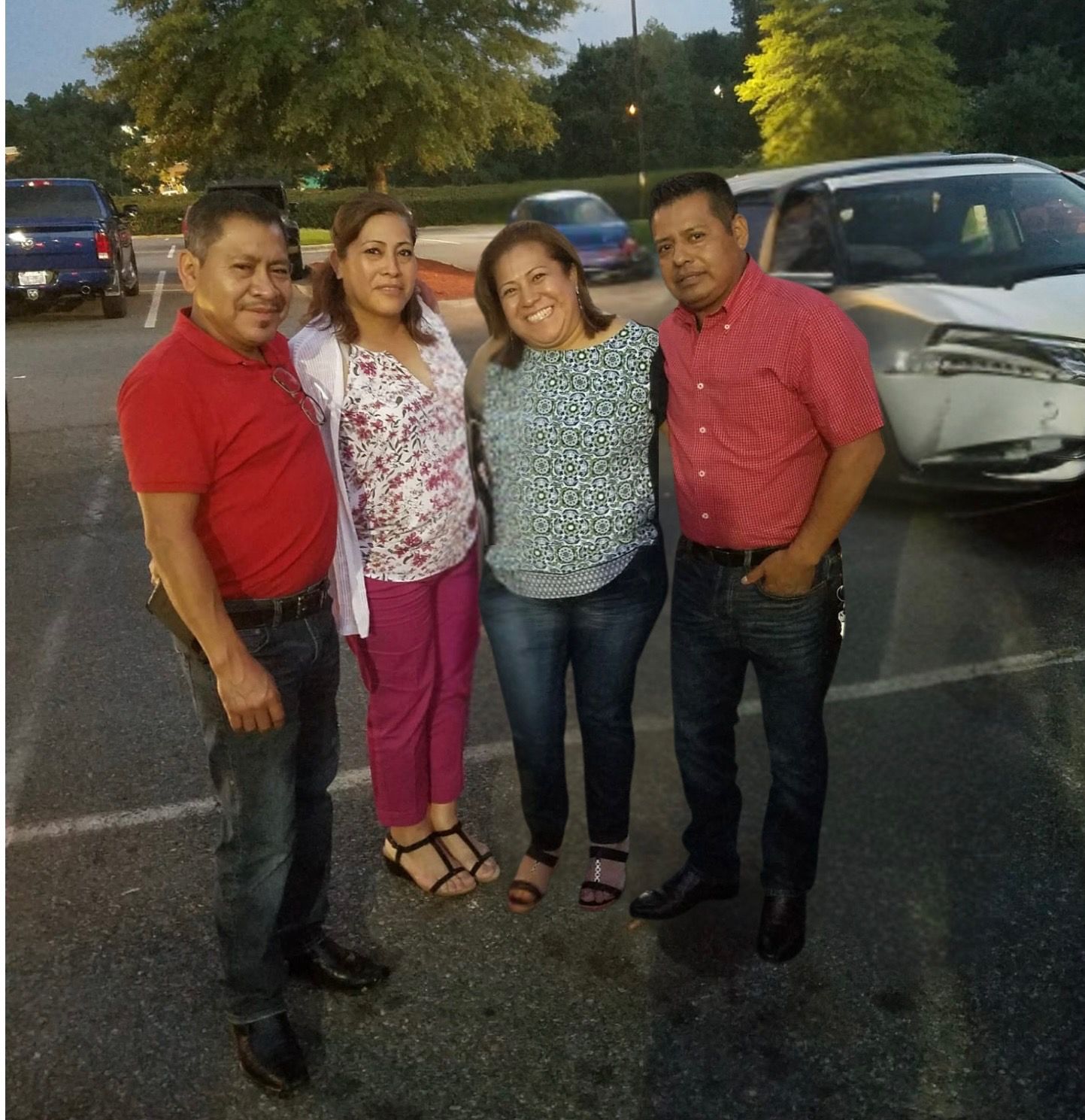 Four adults standing closely together in a parking lot at dusk, two men wearing red shirts and two women in patterned tops, smiling at the camera.