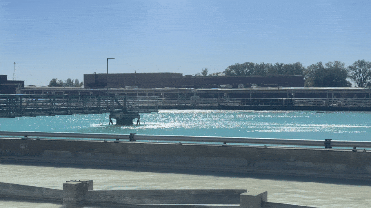 Bright turquoise water with sunlight reflections, surrounded by concrete barriers and metal railings, with industrial buildings and trees in the background under a clear blue sky.