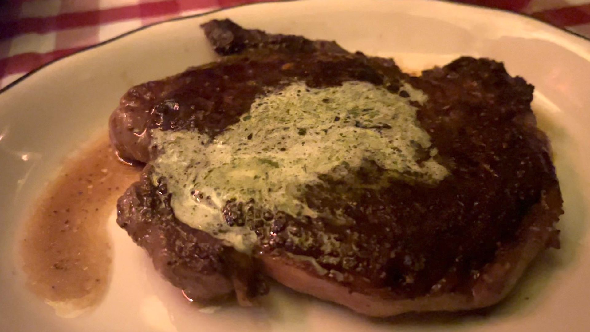 A seared steak on a white plate topped with pale green herb butter, resting in a pool of brown sauce on a red-and-white checkered tablecloth.