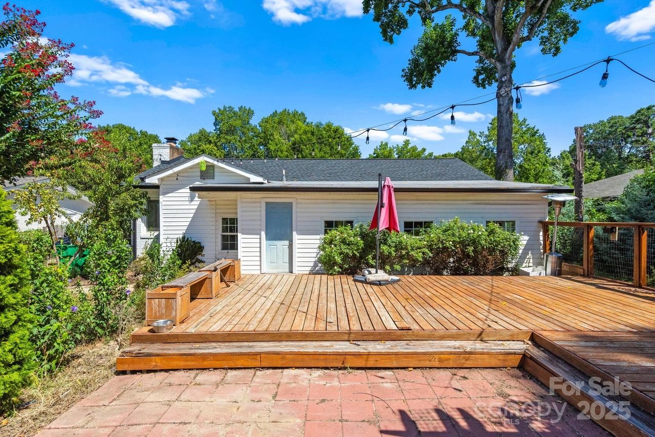 Backyard view of a white house with a large wooden deck, a pink umbrella, string lights overhead, green bushes, and trees under a bright blue sky with scattered clouds.