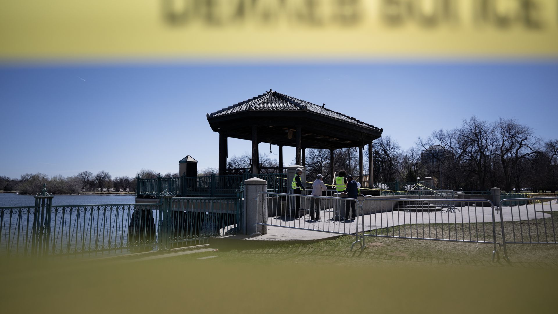 Sunlit lakeside park with a wooden gazebo on stilts over the water. Blurred yellow caution tape frames the foreground as people in high-visibility vests gather by a railing.