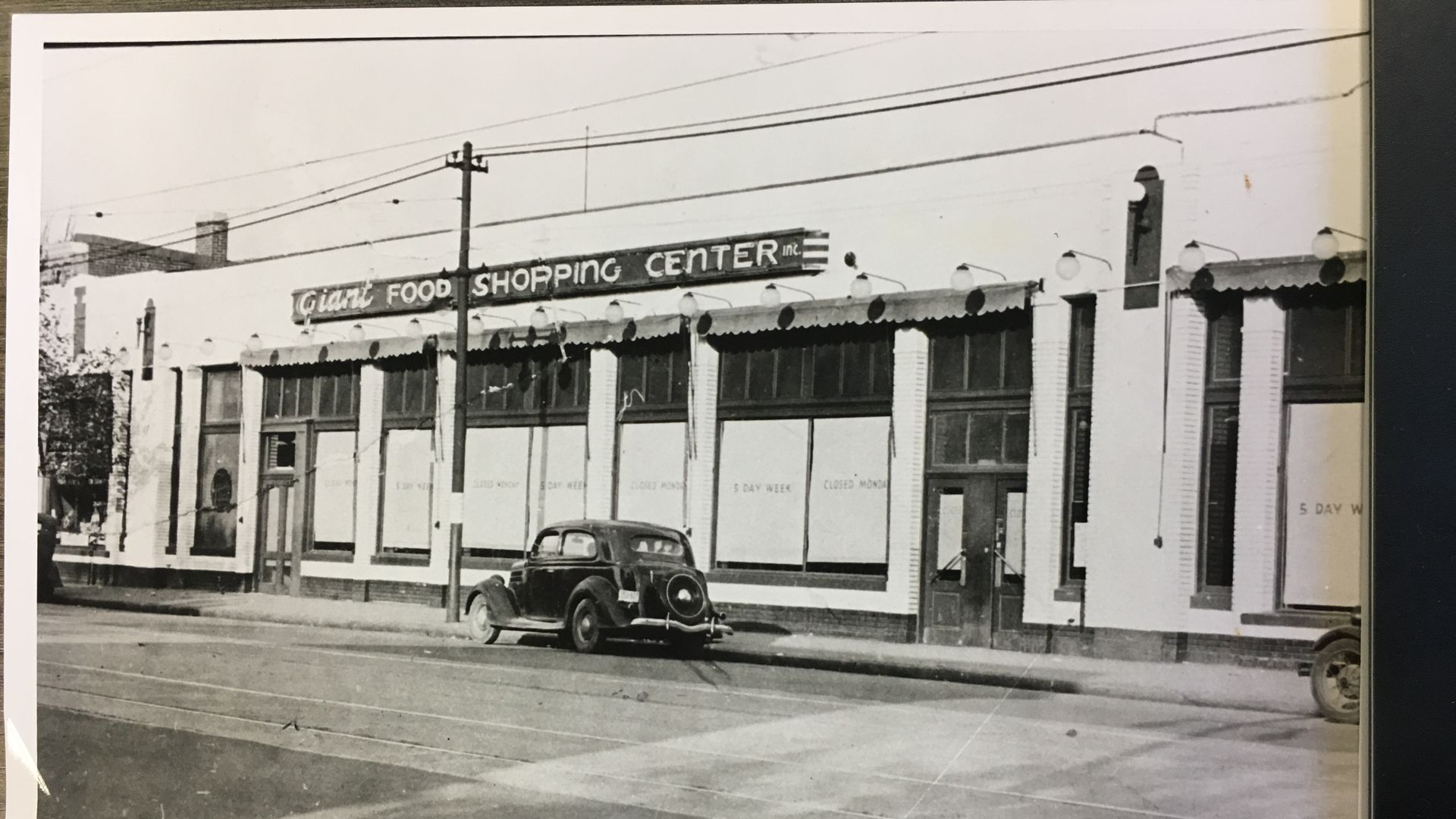 Black and white photo of a vintage car parked in front of a building labeled "Giant Food Shopping Center Inc" with large windows and streetlights above.