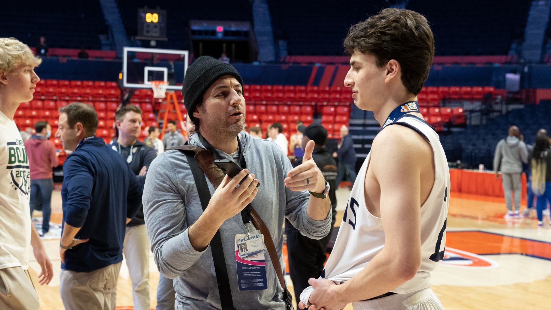 Man in beanie holding a phone interviewing a teenage basketball player on a basketball court.