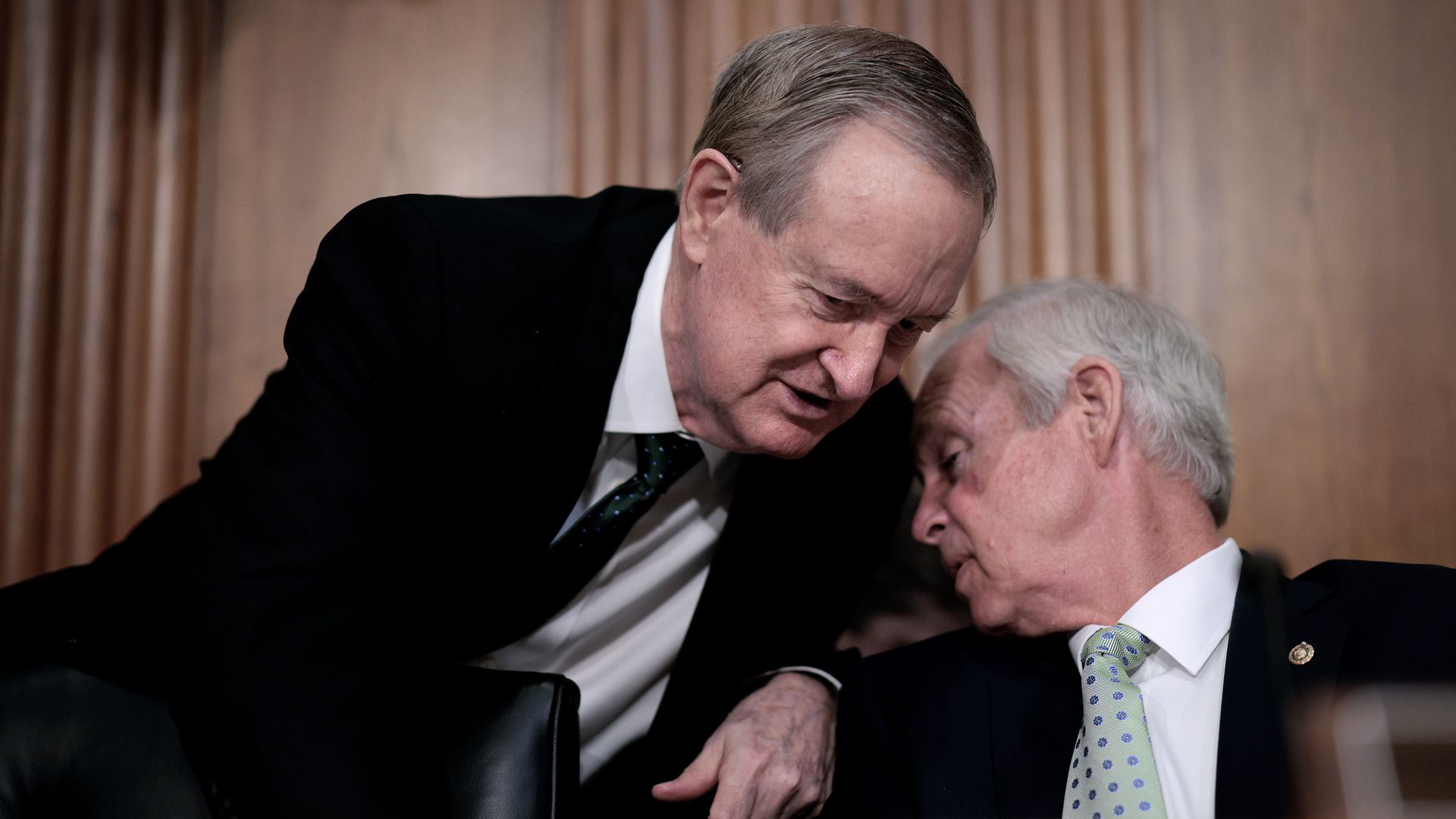 Two men in suits whisper to each other during an official gathering. 