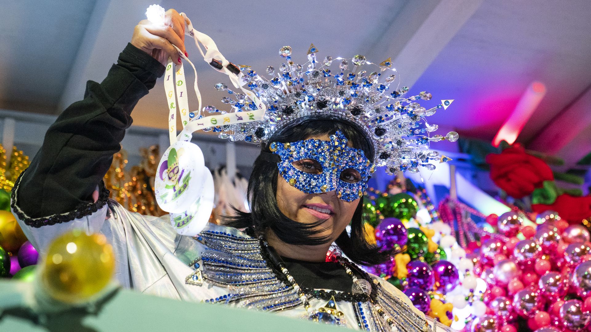 A parade rider, wearing a mask and a decorative headband, tosses beads to a crowd, off-camera.