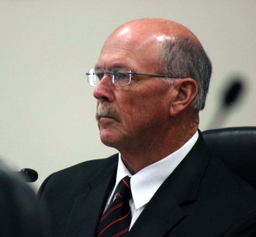 An older man in a black suit with glasses sits in a chair in front of a microphone. 
