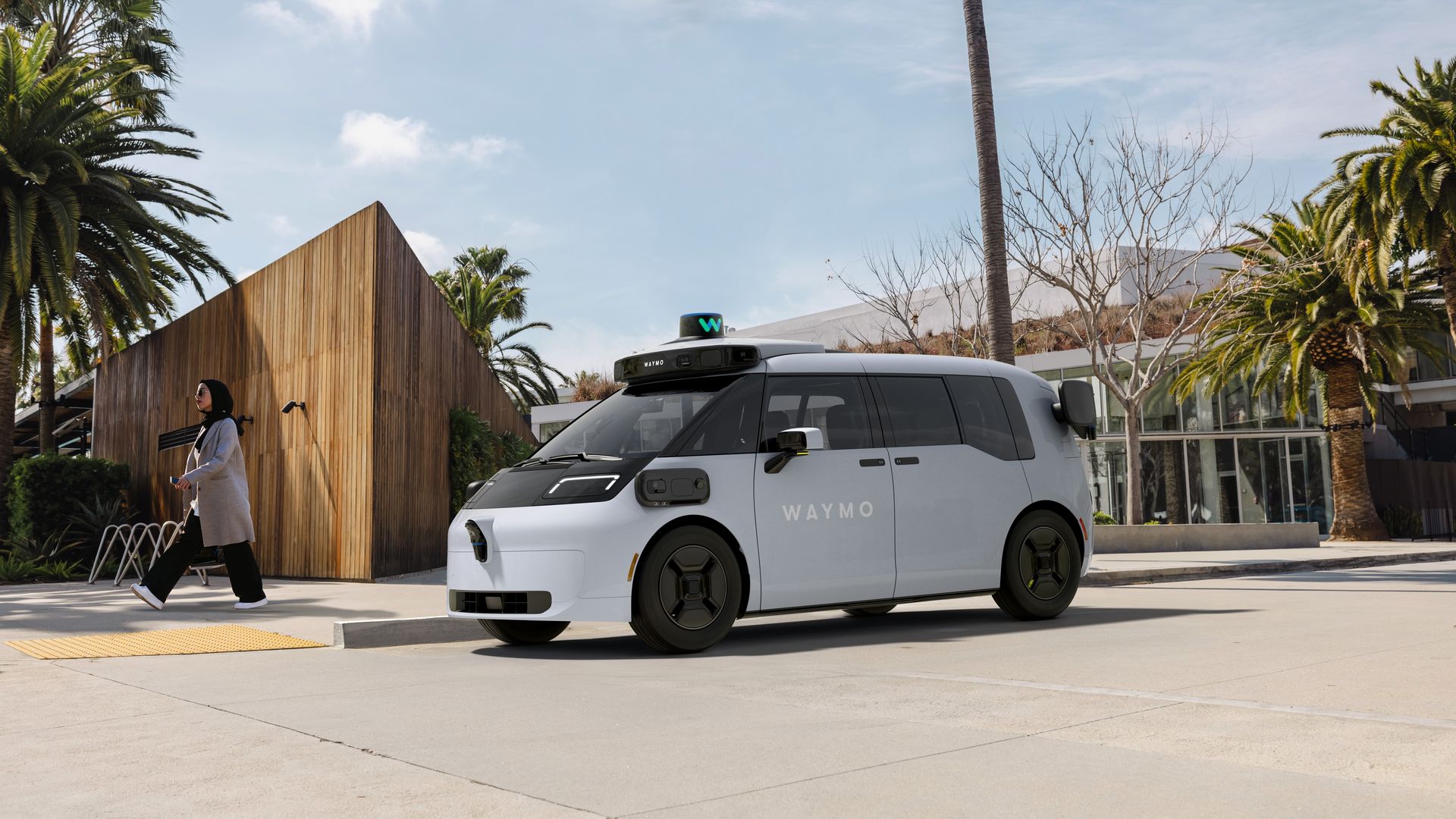 White Waymo autonomous minivan with roof lidar, parked by a wooden wall, palm trees on both sides; a woman wearing a hijab walks past on a sunny day.