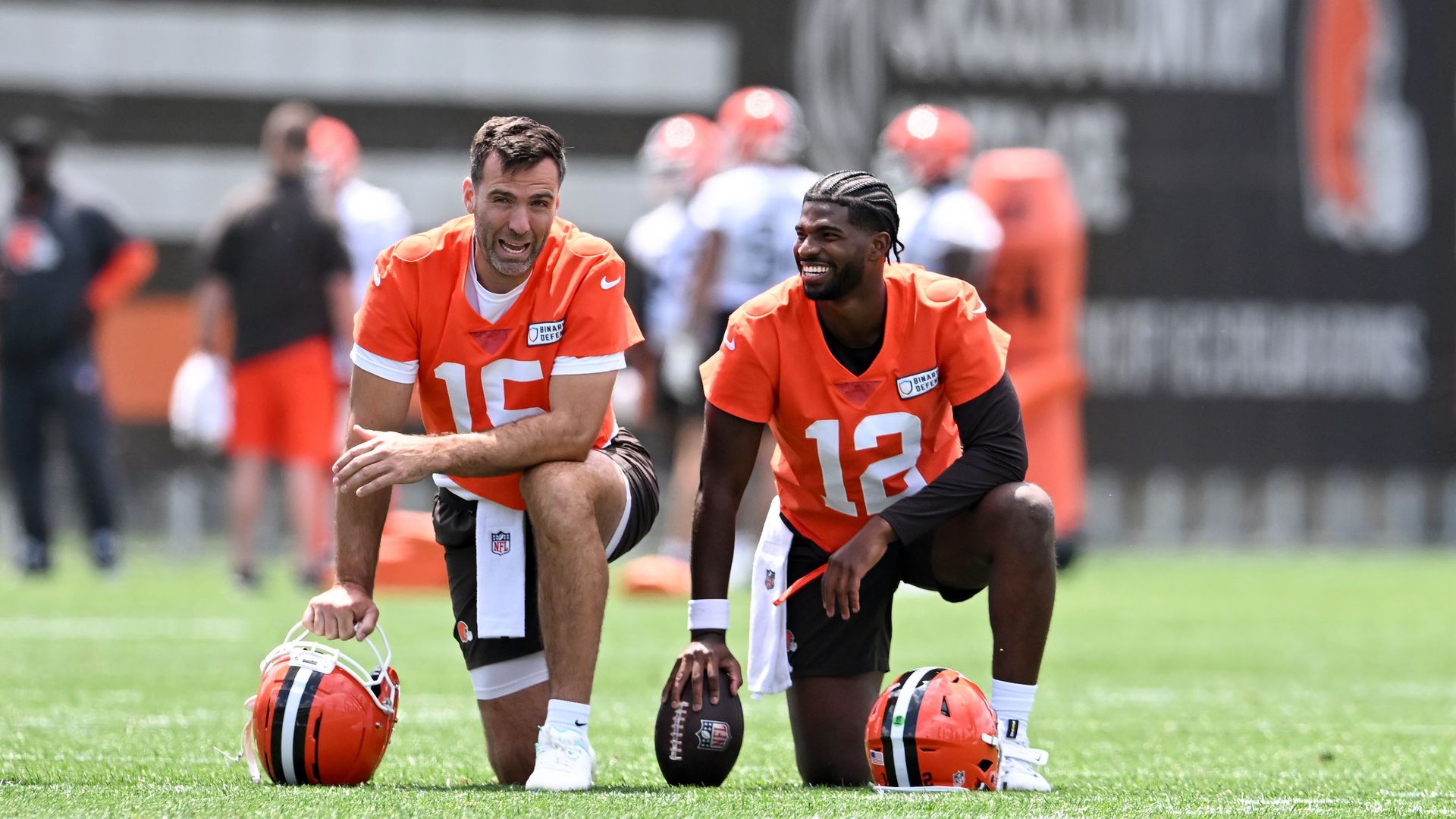 Two football players in orange jerseys numbered 15 and 12 kneel on green field, smiling during practice. Orange helmets rest on ground beside them with blurred teammates and signage in background.