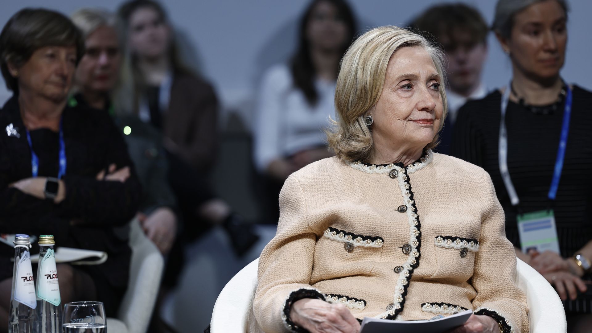 A woman with blonde hair wearing a beige textured jacket with black trim sits in a white chair during an event, holding papers, with a diverse audience blurred in the background.
