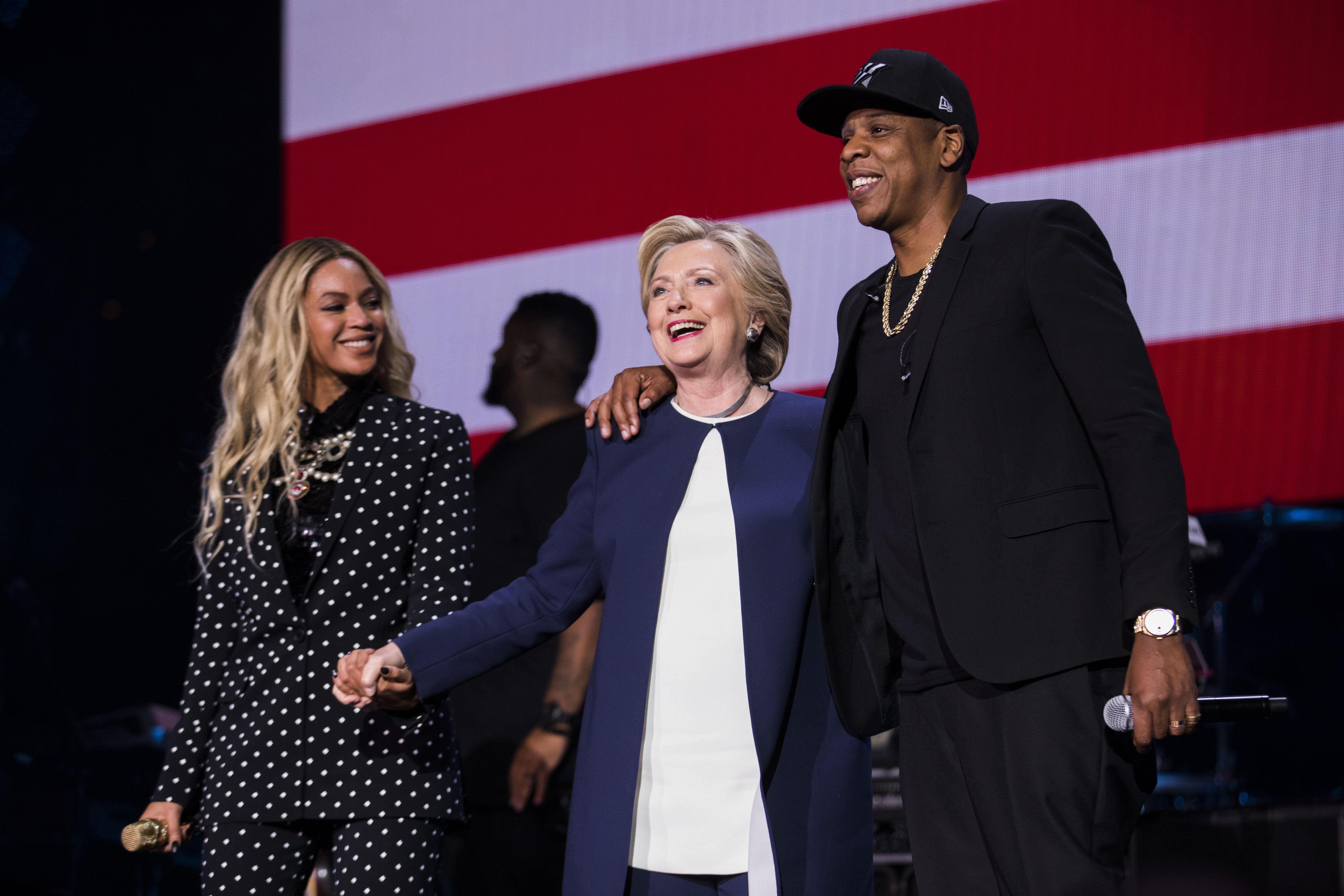 Beyoncé in black and white polka dot suit, Hillary Clinton in navy and white outfit, and Jay-Z in black with gold chain, holding microphones, smiling on stage with an American flag backdrop.