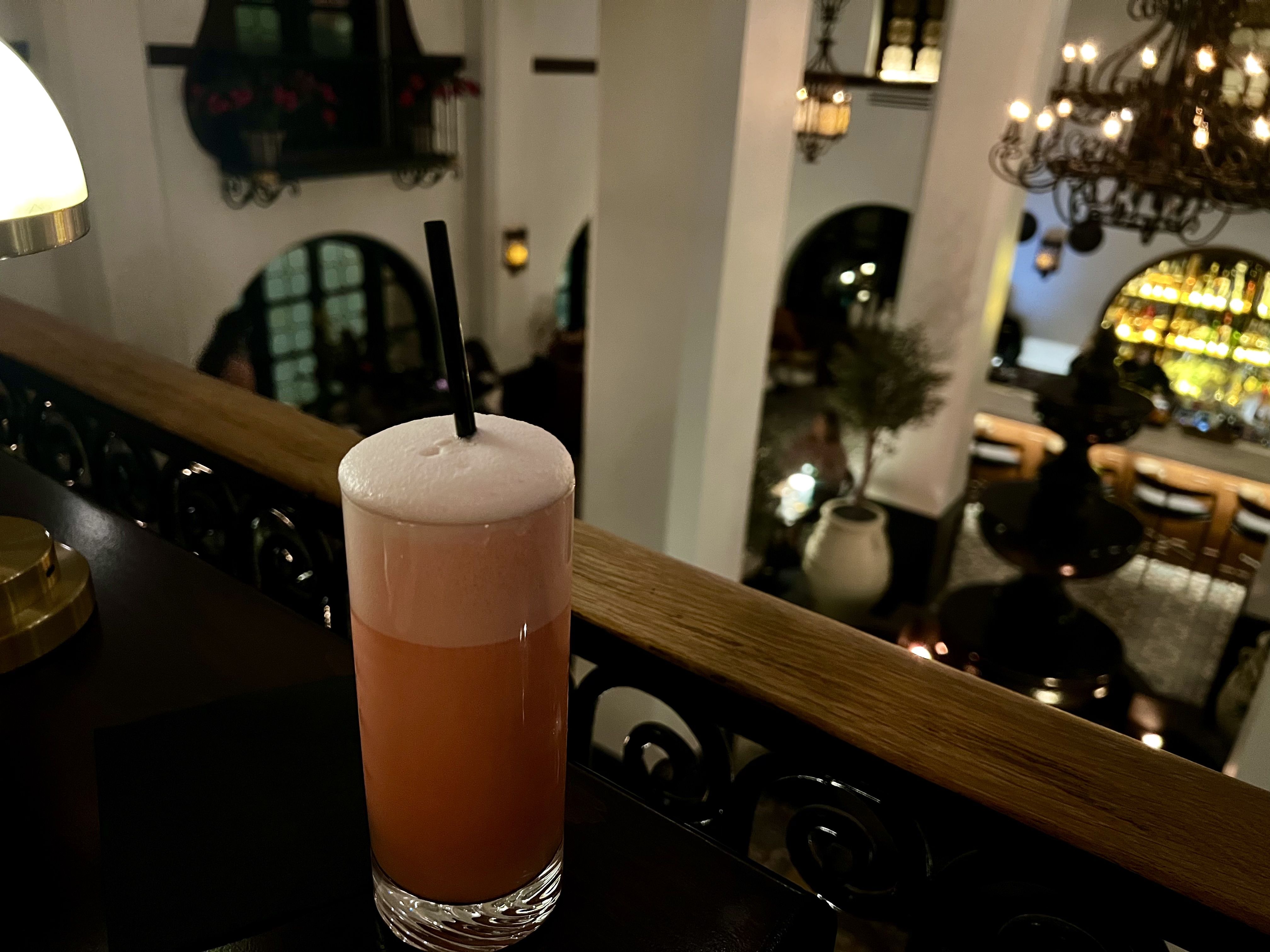Tall glass of pink frothy cocktail with a black straw on a dark bar counter, with a warm-lit elegant bar interior in the background featuring chandeliers and potted plants.