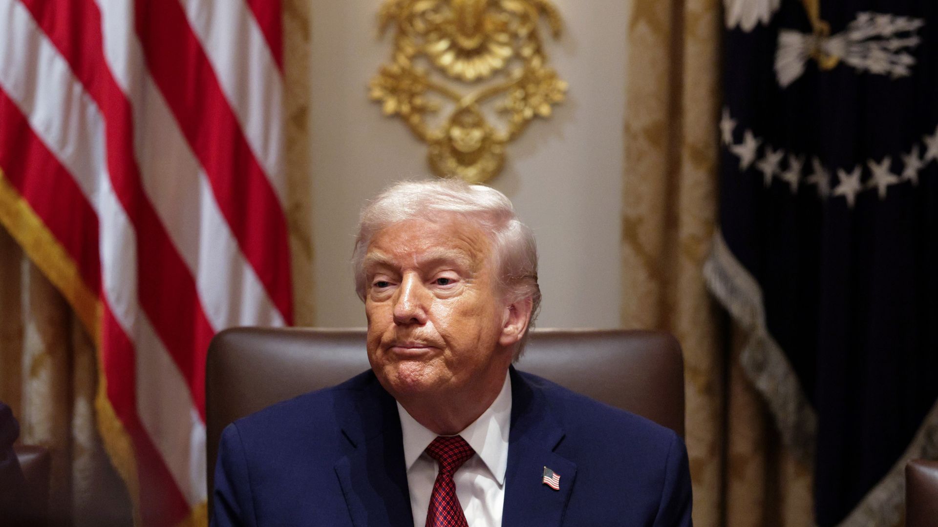 President Trump sits in the Cabinet Room in a brown chair wearing a navy blue suit, a red tie, a white collared shirt and an American flag pin.