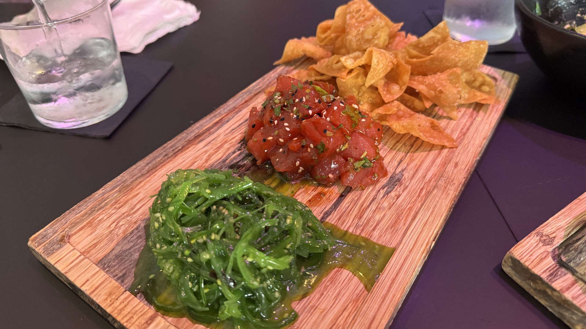 Wooden serving board with a tuna poke of diced red tuna sprinkled with sesame seeds and herbs, a mound of green seaweed salad, and golden tortilla chips on the side; glass of water in the background.