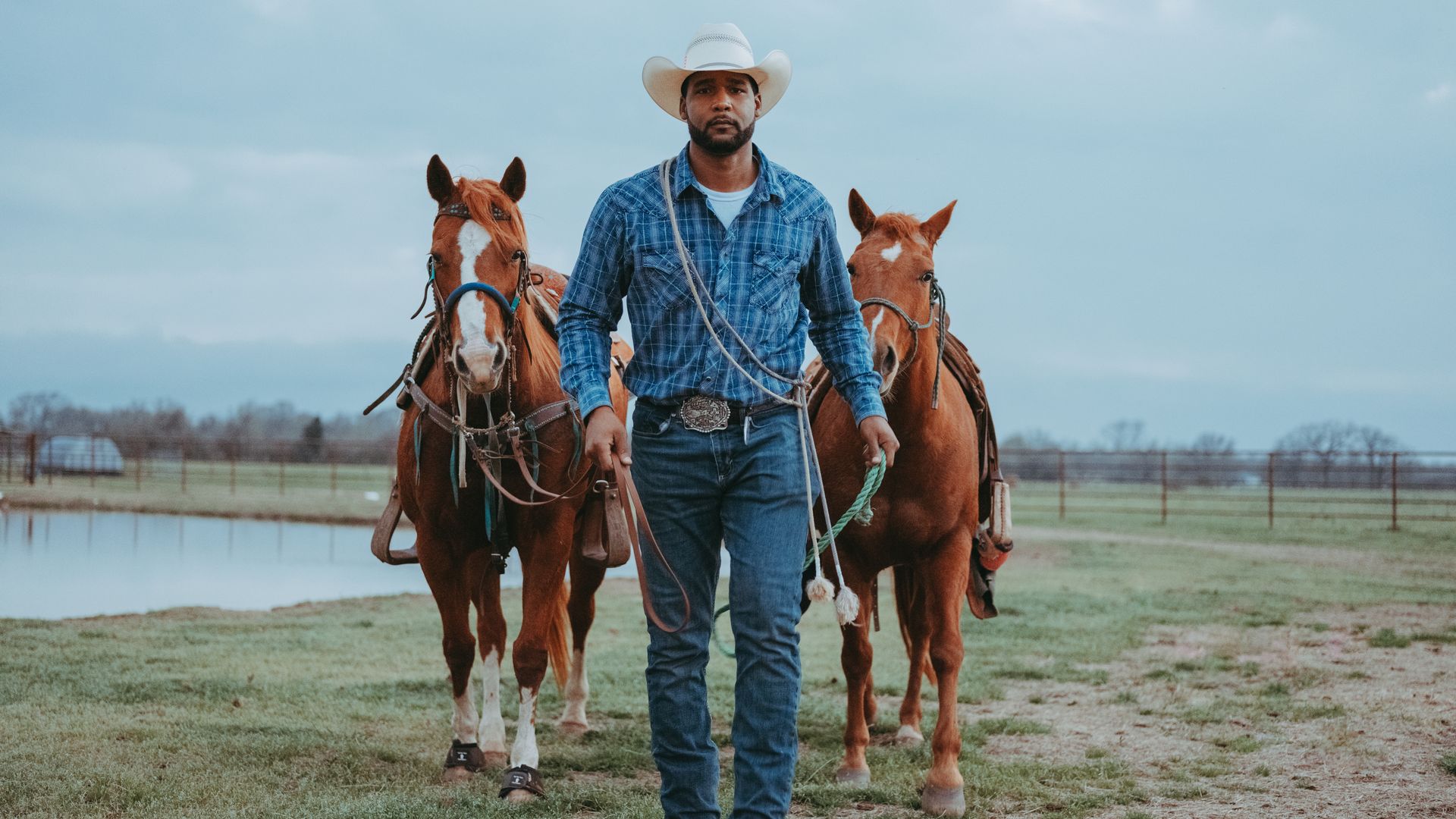 A Black cowboy directing two horses behind him walks straight ahead with a pond behind him.