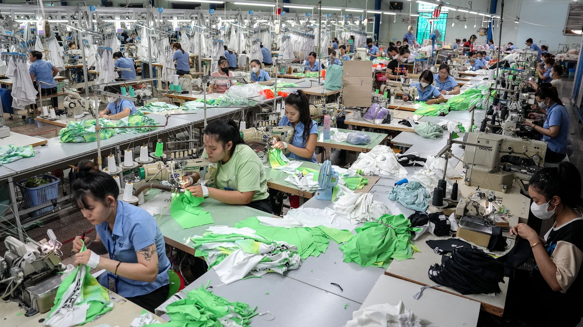 Busy garment factory with many workers at sewing machines stitching bright green and white fabrics; blue uniforms and masks, long rows of workstations under fluorescent lights.