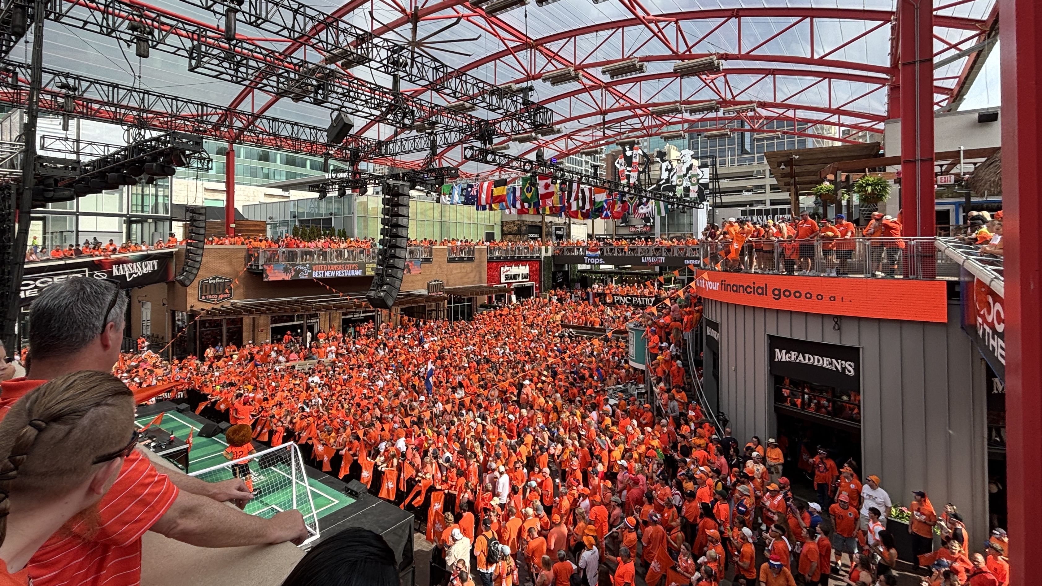 Wide view of KC Live! filled with orange-clad fans; a red steel rig of lights and speakers spans the ceiling; flags hang between balconies while spectators fill the lower levels and mezzanines.