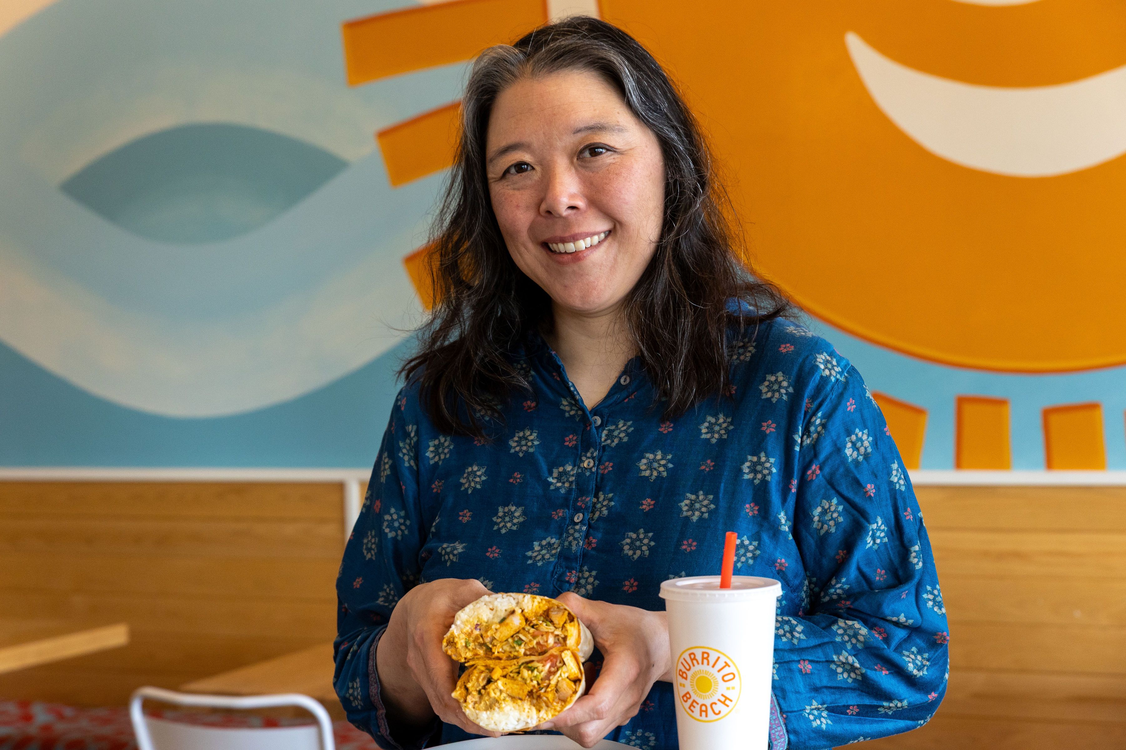 Smiling woman with dark hair and a blue floral shirt holding a burrito cut in half, sitting at a table with a white cup labeled "Burrito Beach" and colorful mural background.