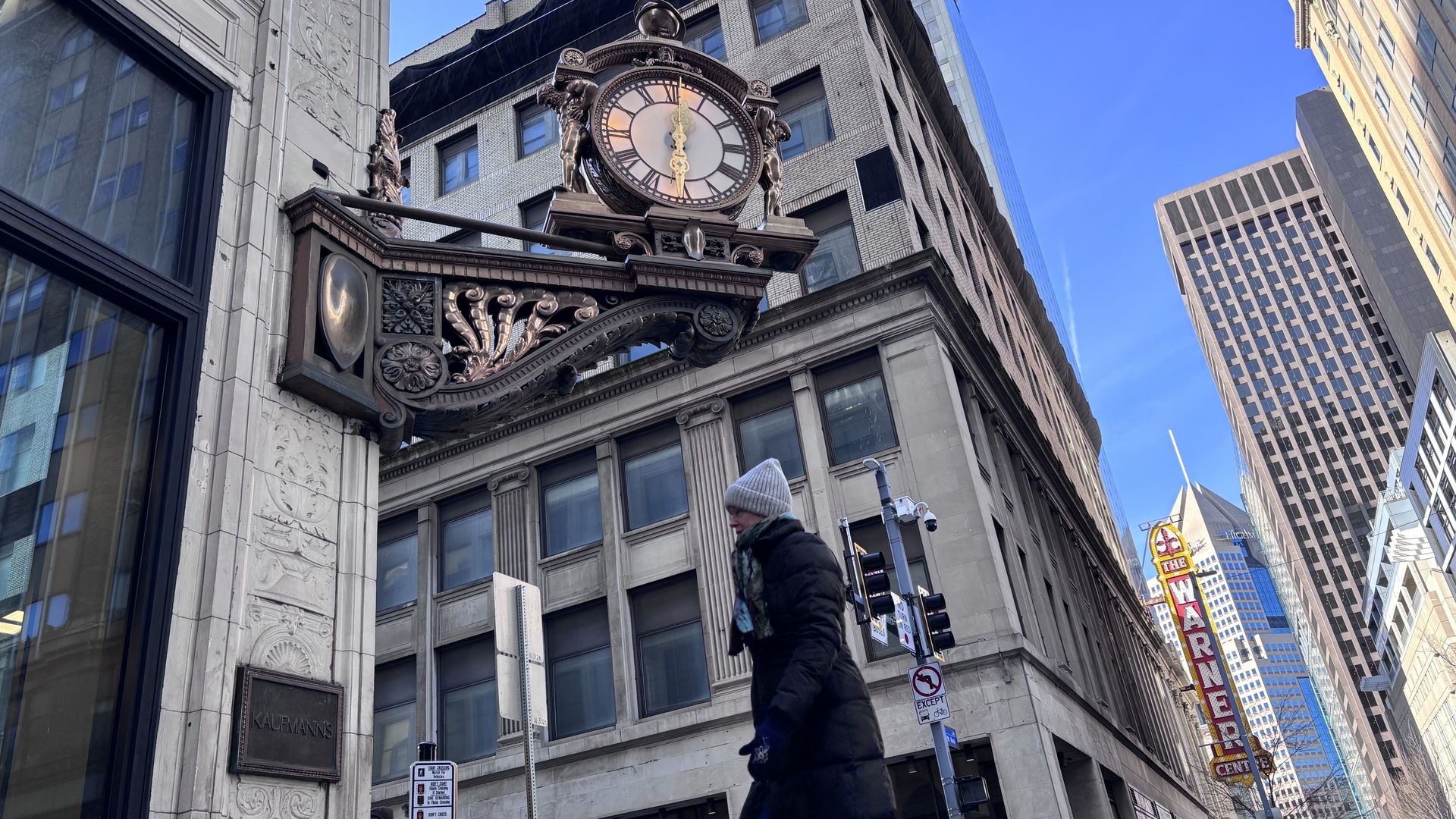 Ornate clock on corner of Kaufmann's building in city with tall buildings, blue sky, and a person in winter clothes walking by.