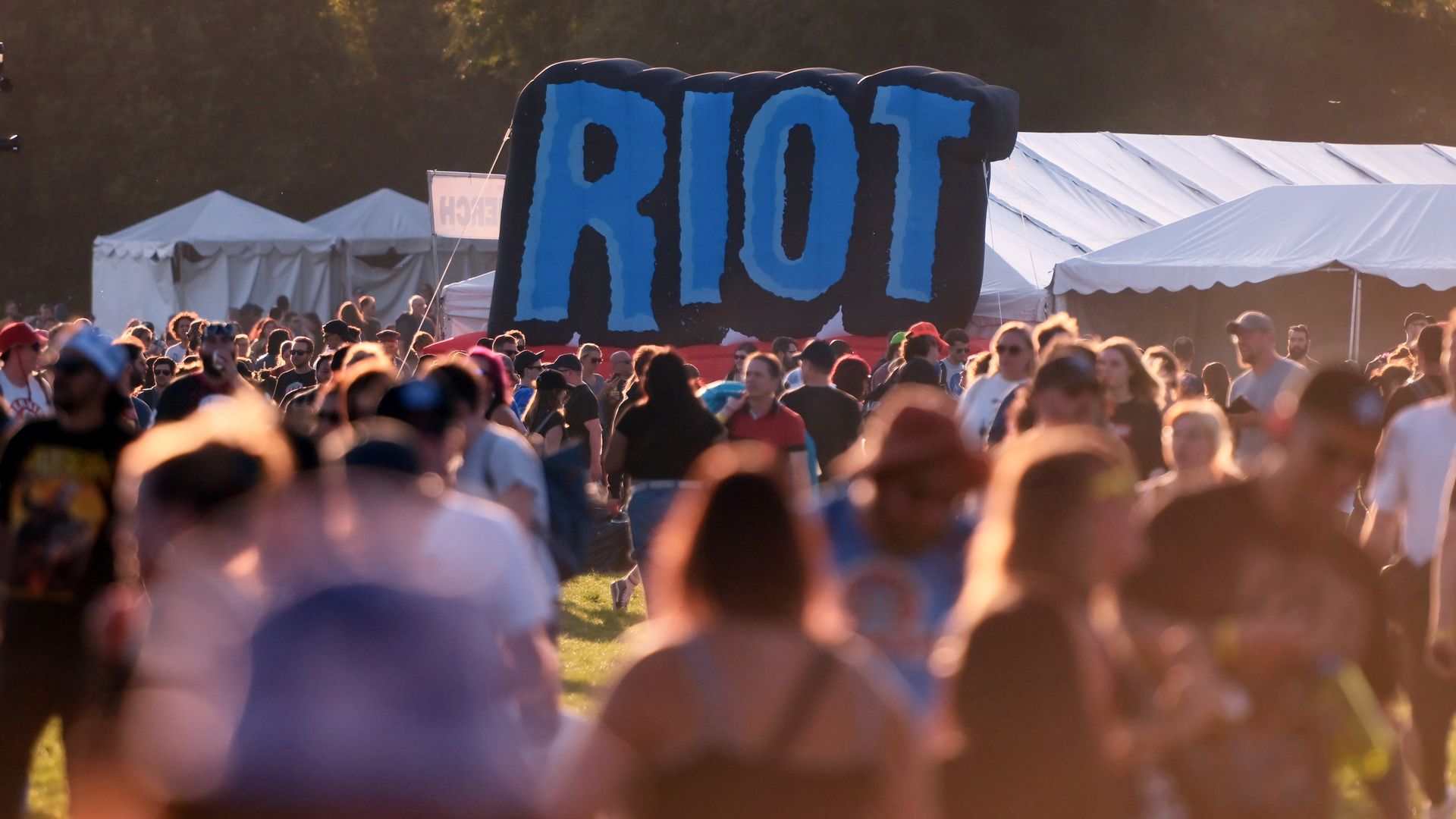 Large blue letters on a black inflatable sign rise above a crowded outdoor festival, with white tents in the background and warm sunset light.