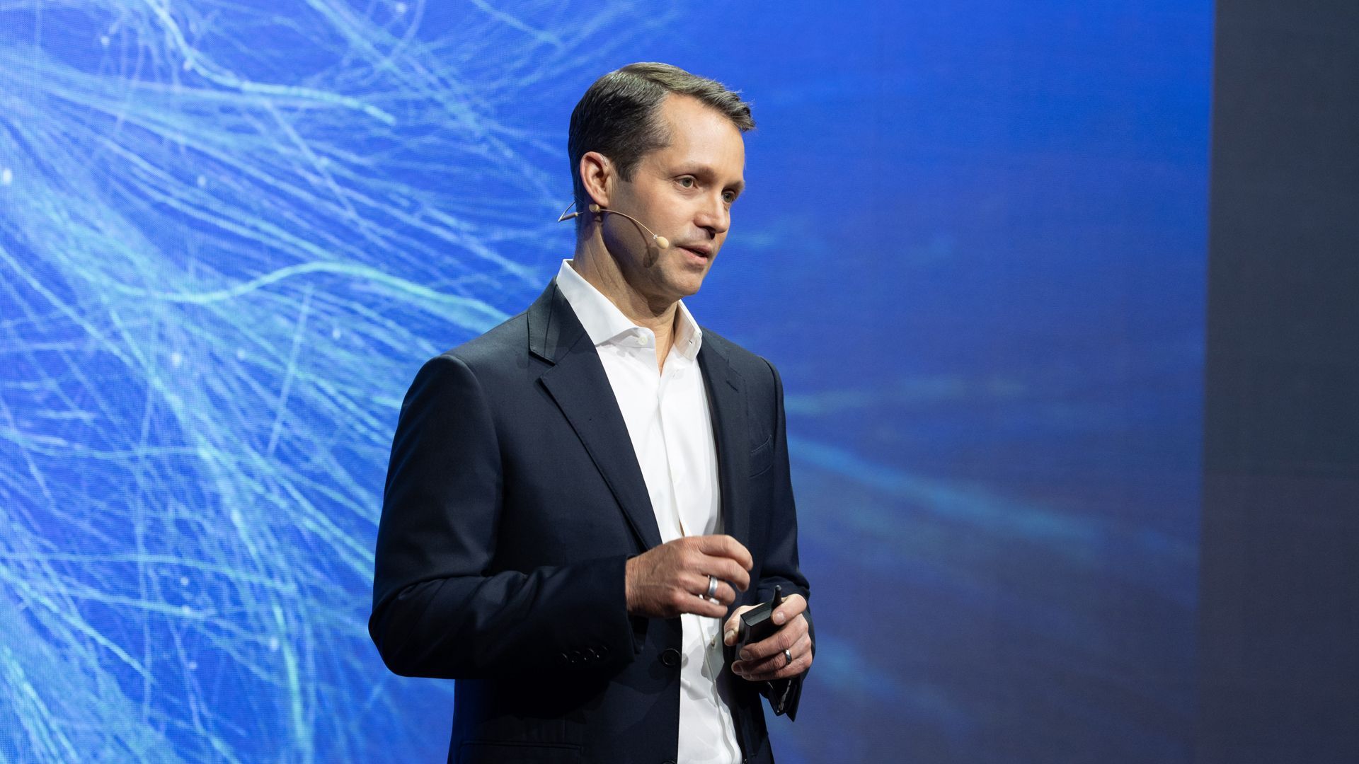 A man in a dark suit and white shirt speaks on stage with a wireless headset microphone, in front of a blue digital network background.