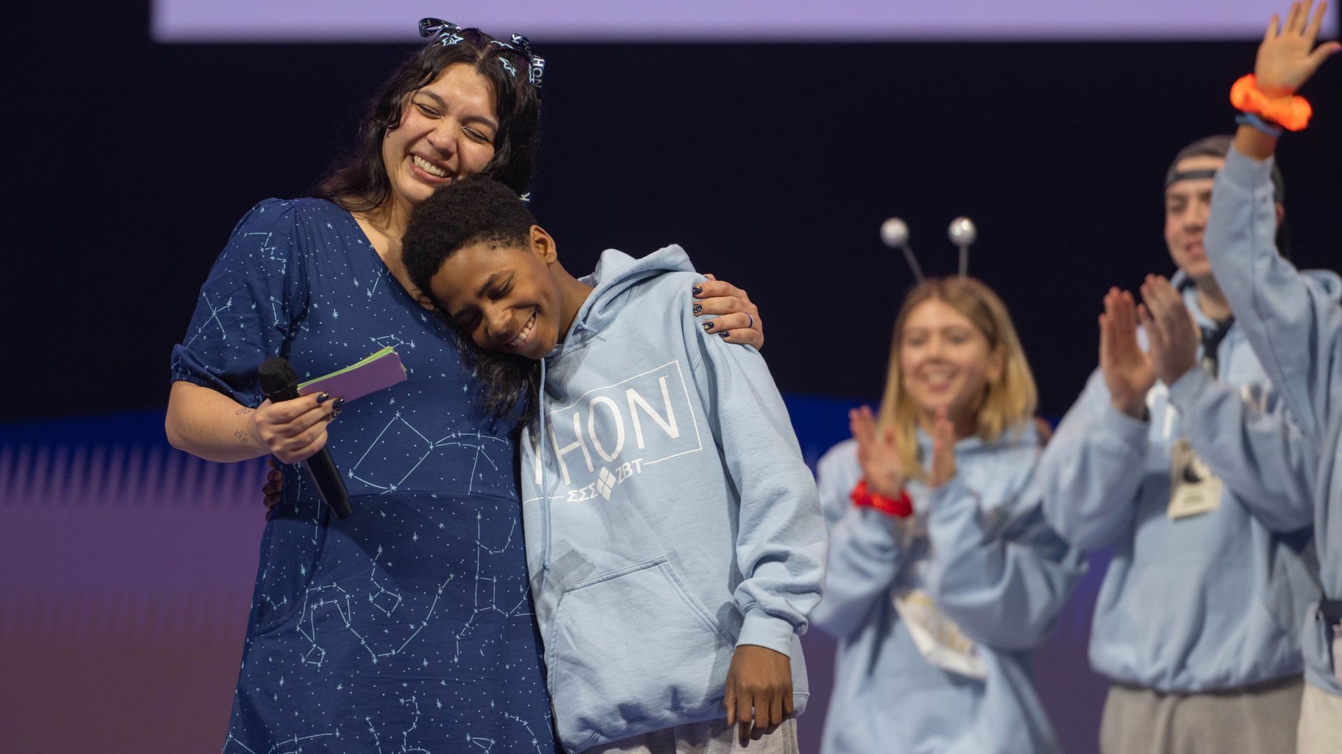 A woman in a dark blue constellation-patterned dress embraces and smiles with a boy in a light blue hoodie labeled "THON" on stage, while two people in blue hoodies applaud in the background.
