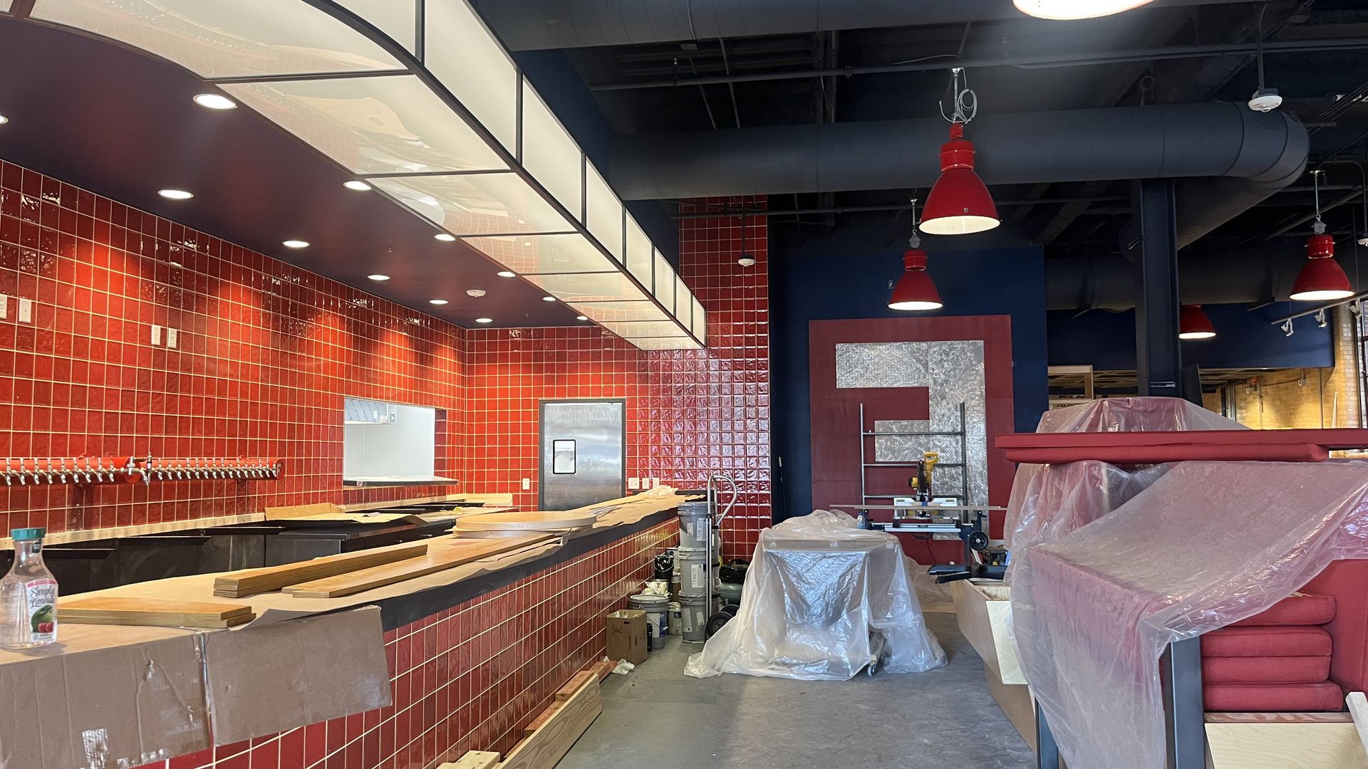 Brewery under renovation with glossy red tile walls, a long bar wrapped in plastic, and exposed blue ductwork. Red pendant lights hang from the ceiling amid construction materials.