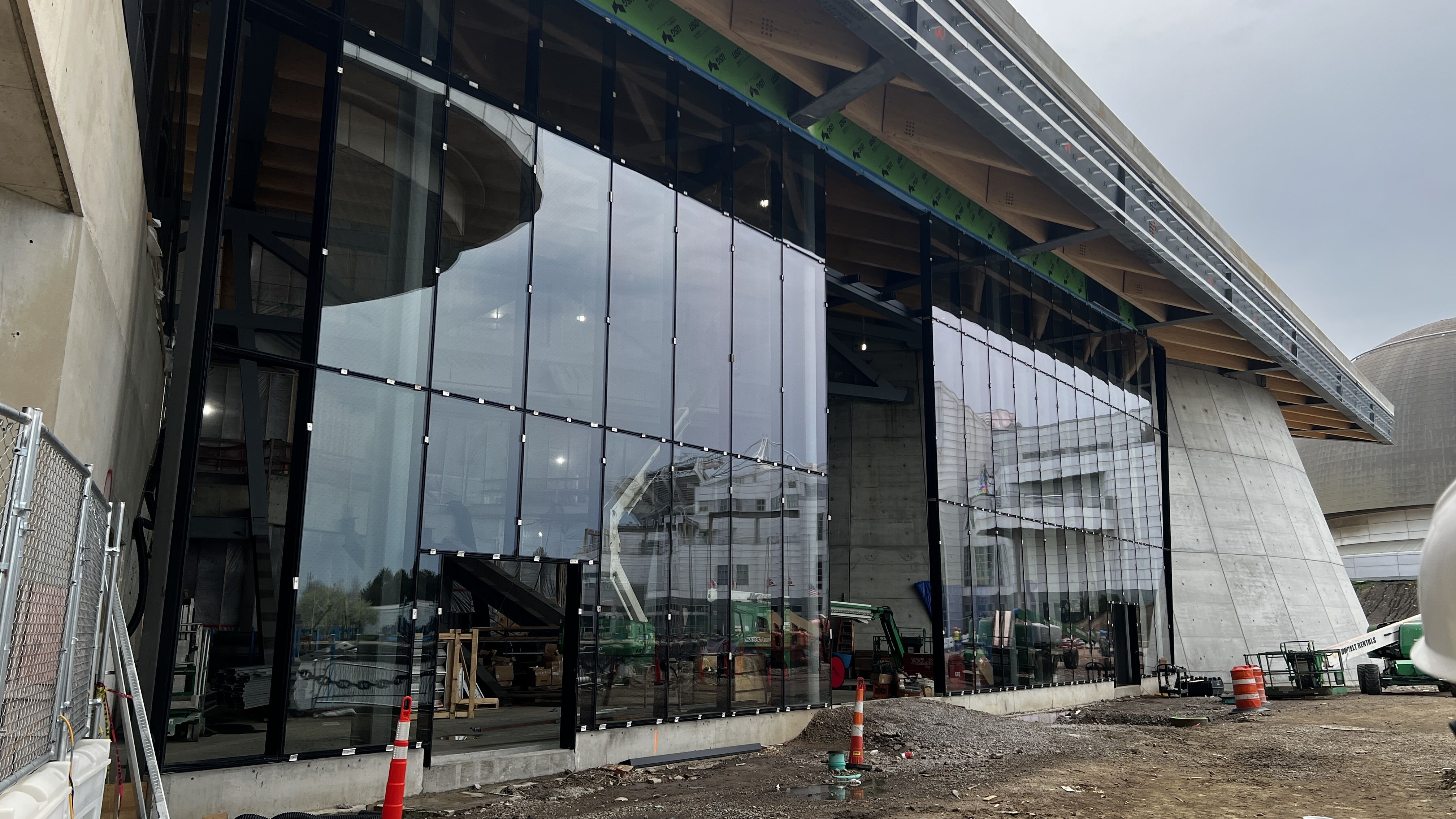 Modern glass storefront under construction with tall black steel frames and large reflective panels along a concrete base. Green wrap on upper beam; construction equipment and orange cones nearby.