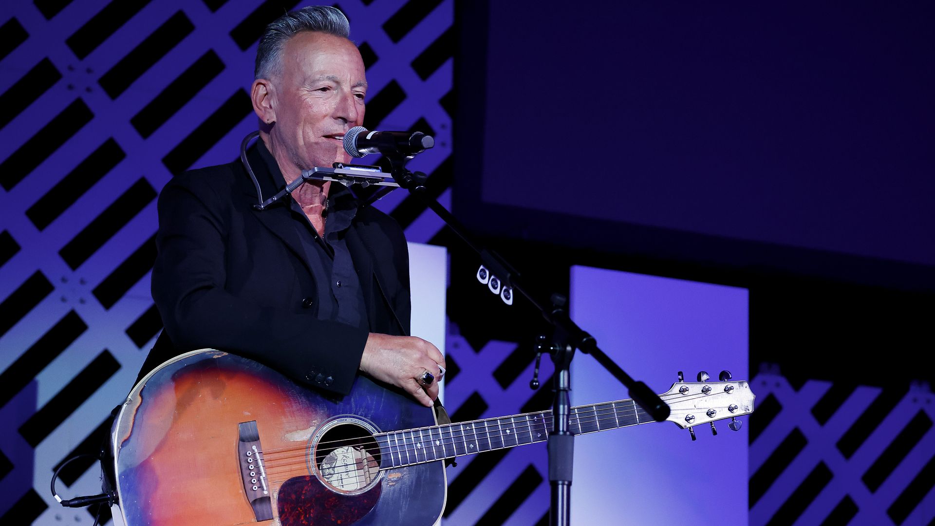 A gray-haired man in a dark jacket plays a sunburst acoustic guitar and sings into a microphone on a stage with purple lighting and a lattice backdrop.