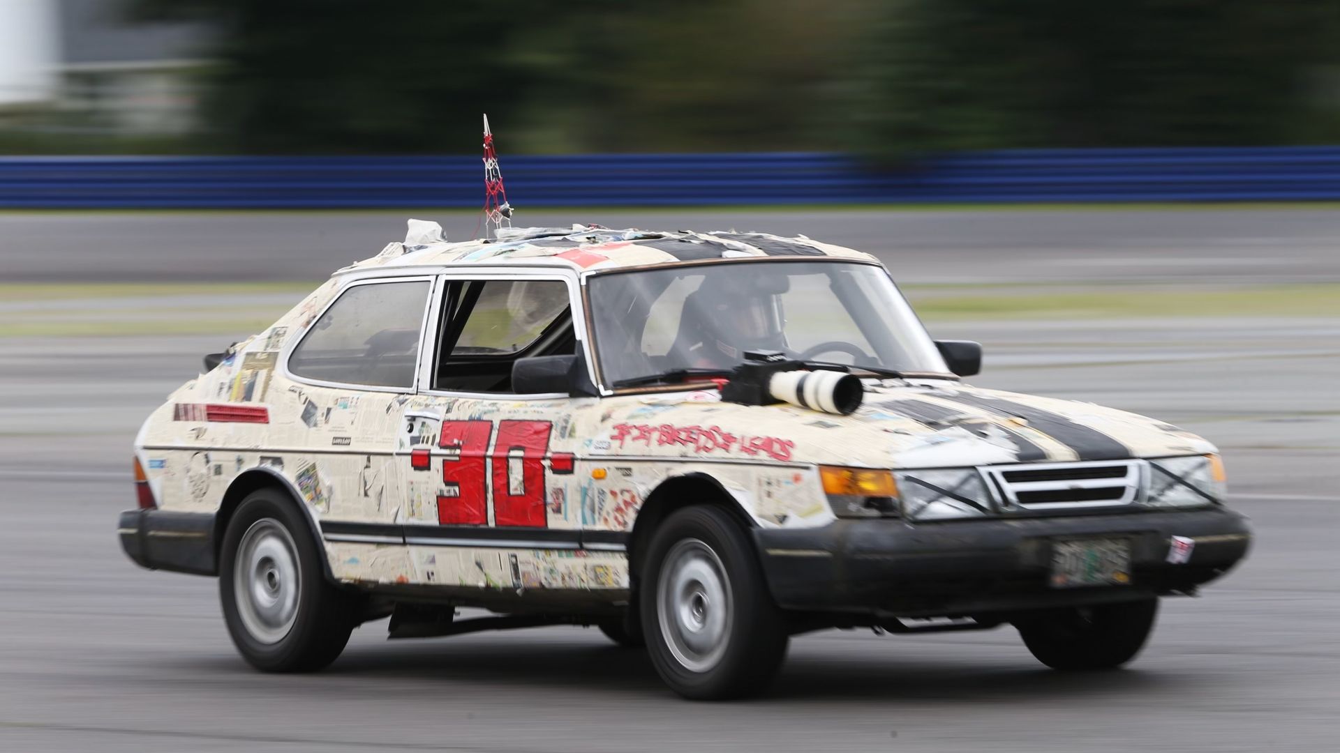 White car covered in newspaper print with large red number 30 on the side, speeding on a race track with a camera lens mounted on the hood; driver wearing helmet visible inside.