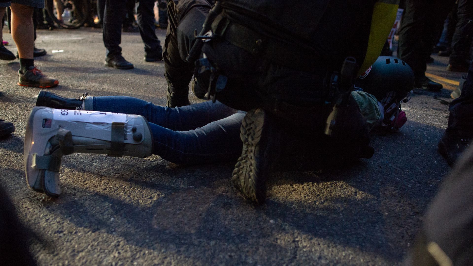 Dim street scene with a person in boots lying on the pavement, surrounded by a crowd. Tactical gear, a large backpack, rope, and a helmet sit nearby as others stand.