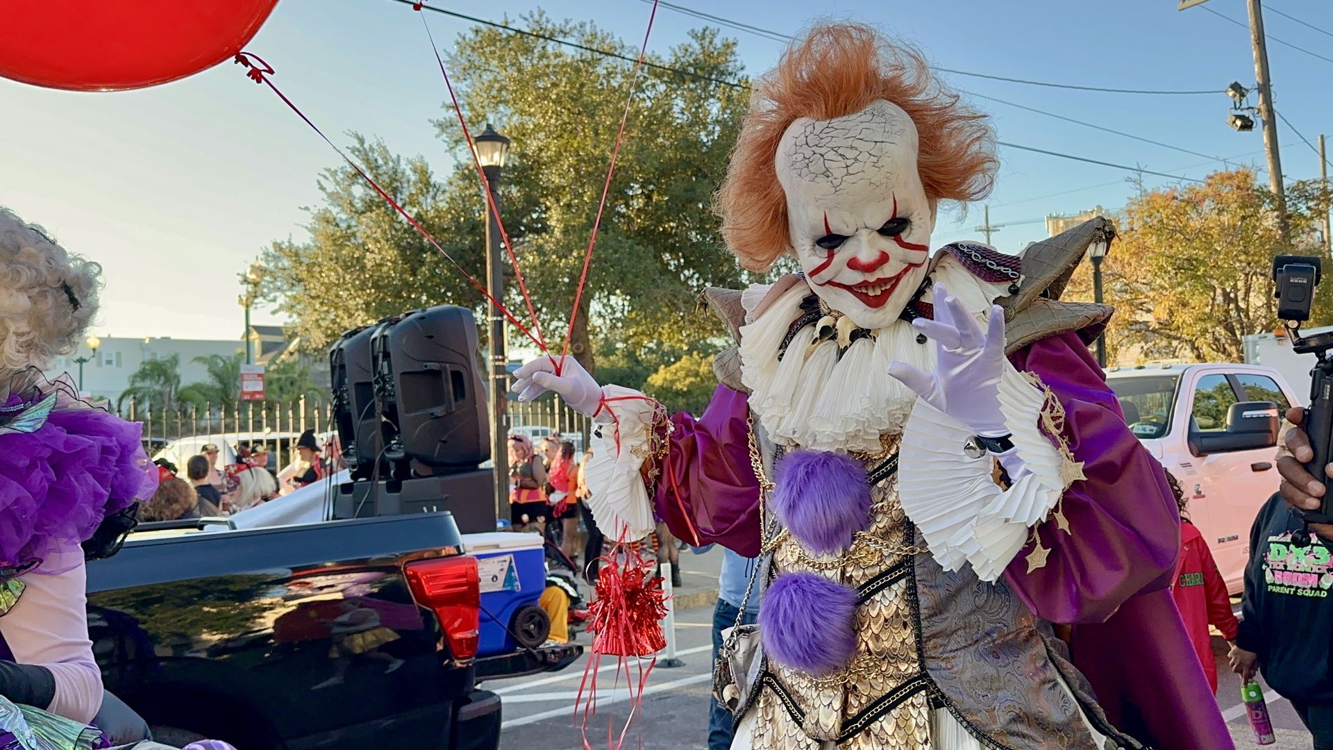 Costumed crowd scene with a sinister white-faced clown, orange hair, red makeup, and a purple-gold outfit, holding red balloon strings during a street parade; onlookers and a photographer nearby.