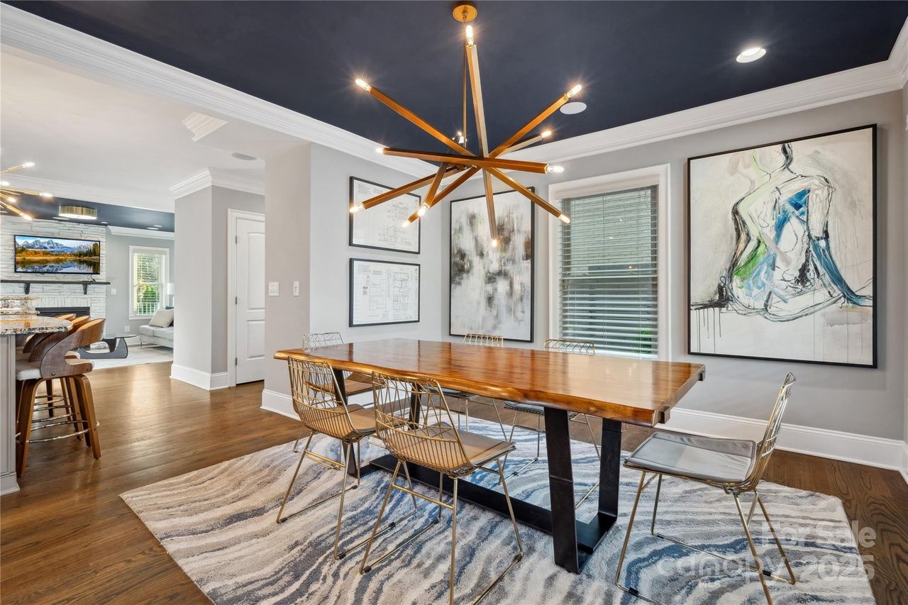 Modern dining room with wooden table, metal chairs, blue and white geometric rug, dark blue ceiling, abstract art on walls, and unique starburst chandelier lighting.