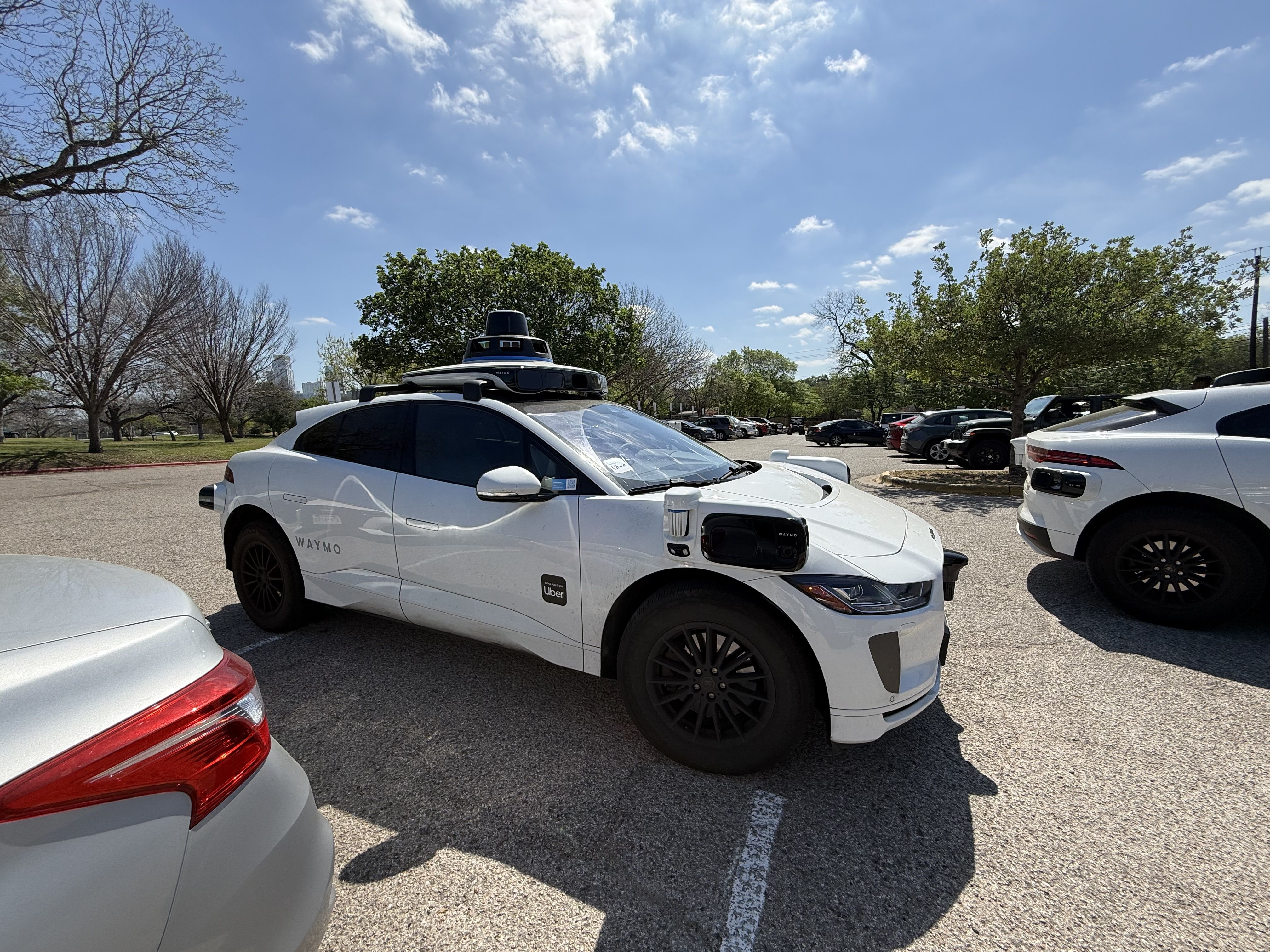 White Waymo self-driving car with roof-mounted lidar, Uber sticker, parked in a sunny parking lot among other cars, with trees and a blue sky in the background.