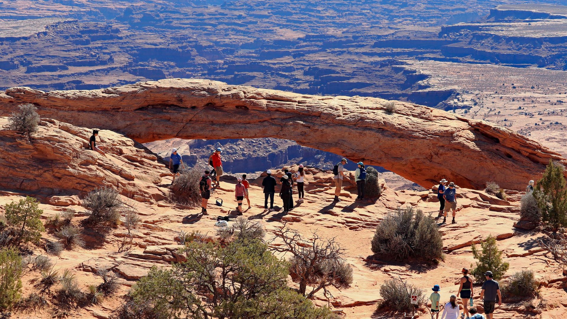Group of hikers under a large red sandstone arch in a desert canyon, with layered canyon walls in the distance and sparse vegetation in the foreground.