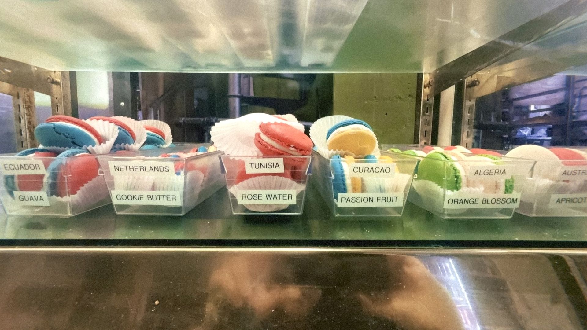 Colorful macarons in clear plastic cups on a metal shelf, labeled by country: Ecuador (guava), Netherlands (cookie butter), Tunisia (rose water), Curacao (passion fruit), Algeria (orange blossom).