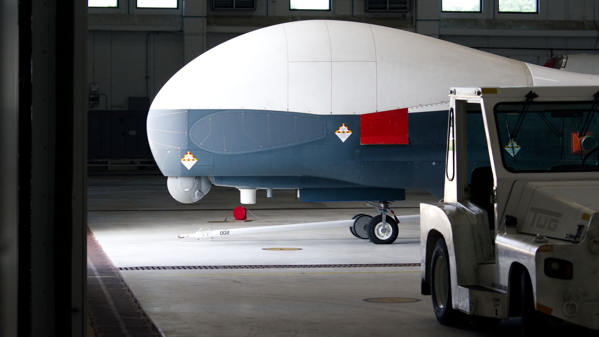 Inside a hangar, a large white and grey aircraft fuselage with a red panel sits on the tarmac. A tow tractor is nearby and a long rod lies on the floor.