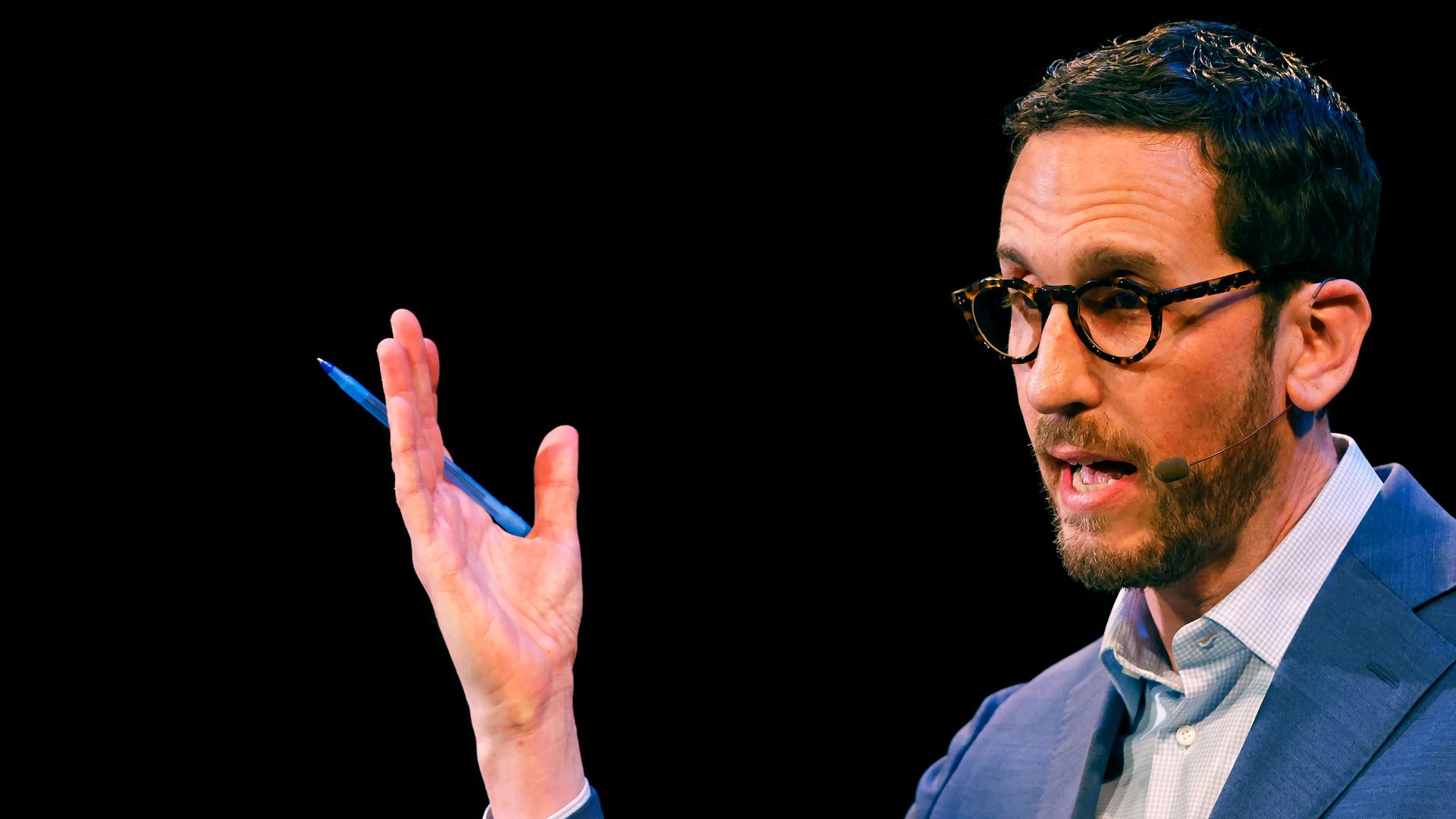 A man with dark hair and a short beard, wearing tortoiseshell glasses, a blue suit and light shirt, speaks into a headset microphone while gesturing with a blue pen against a black background.