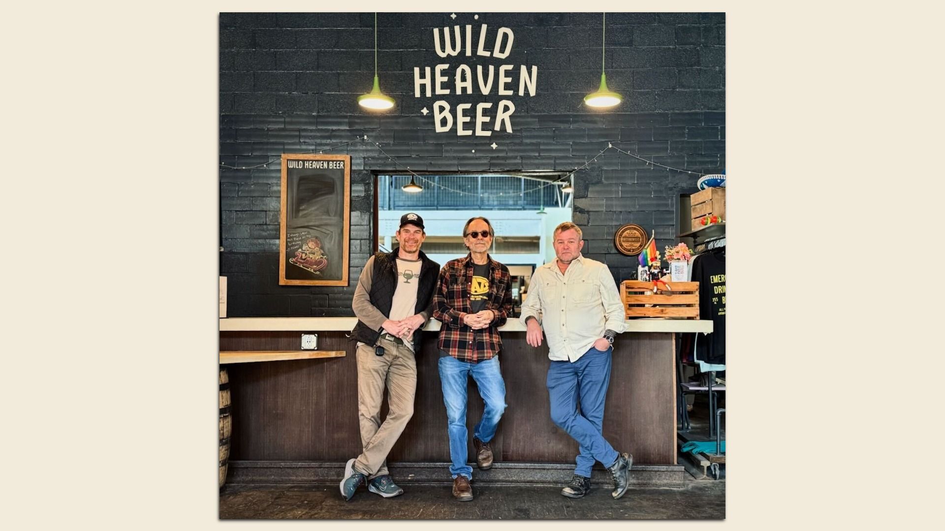 Three men standing in front of a bar with a black brick wall behind them displaying the words "WILD HEAVEN BEER" in white letters, two hanging lights above, and various decorations on the bar.