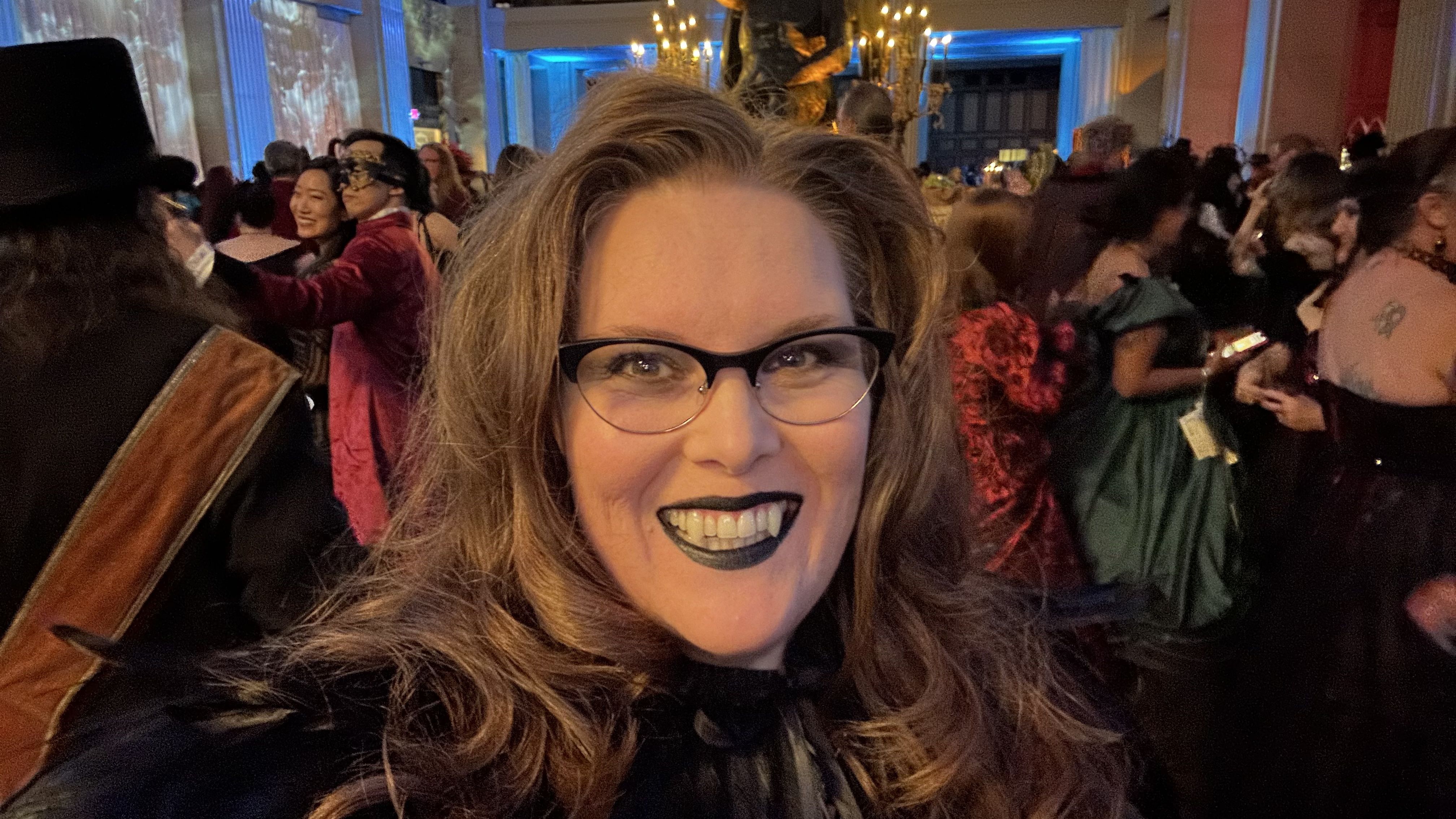 Smiling woman with long hair, black lipstick, and glasses in a crowded room with people in colorful costumes and masks, warm lighting, and blue-lit walls in the background.