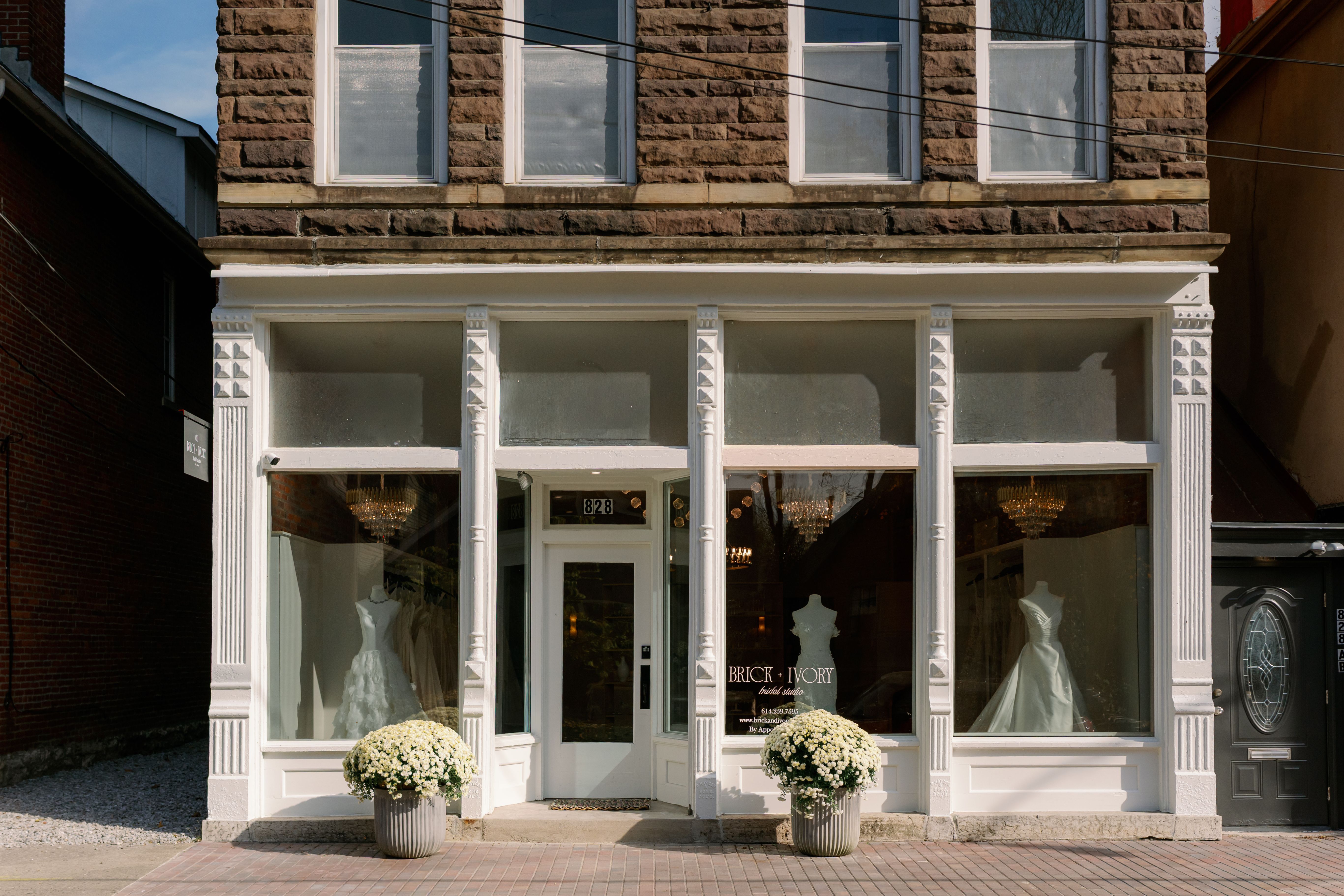 The new Brick + Ivory bridal studio storefront with three large display windows showing wedding dresses on mannequins, two large white flower pots, and a white framed glass door.