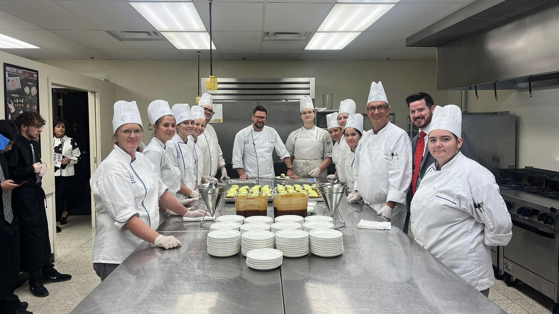 Group of chefs and culinary students wearing white uniforms and hats standing around a stainless steel kitchen table with plates and trays of food in a professional kitchen.