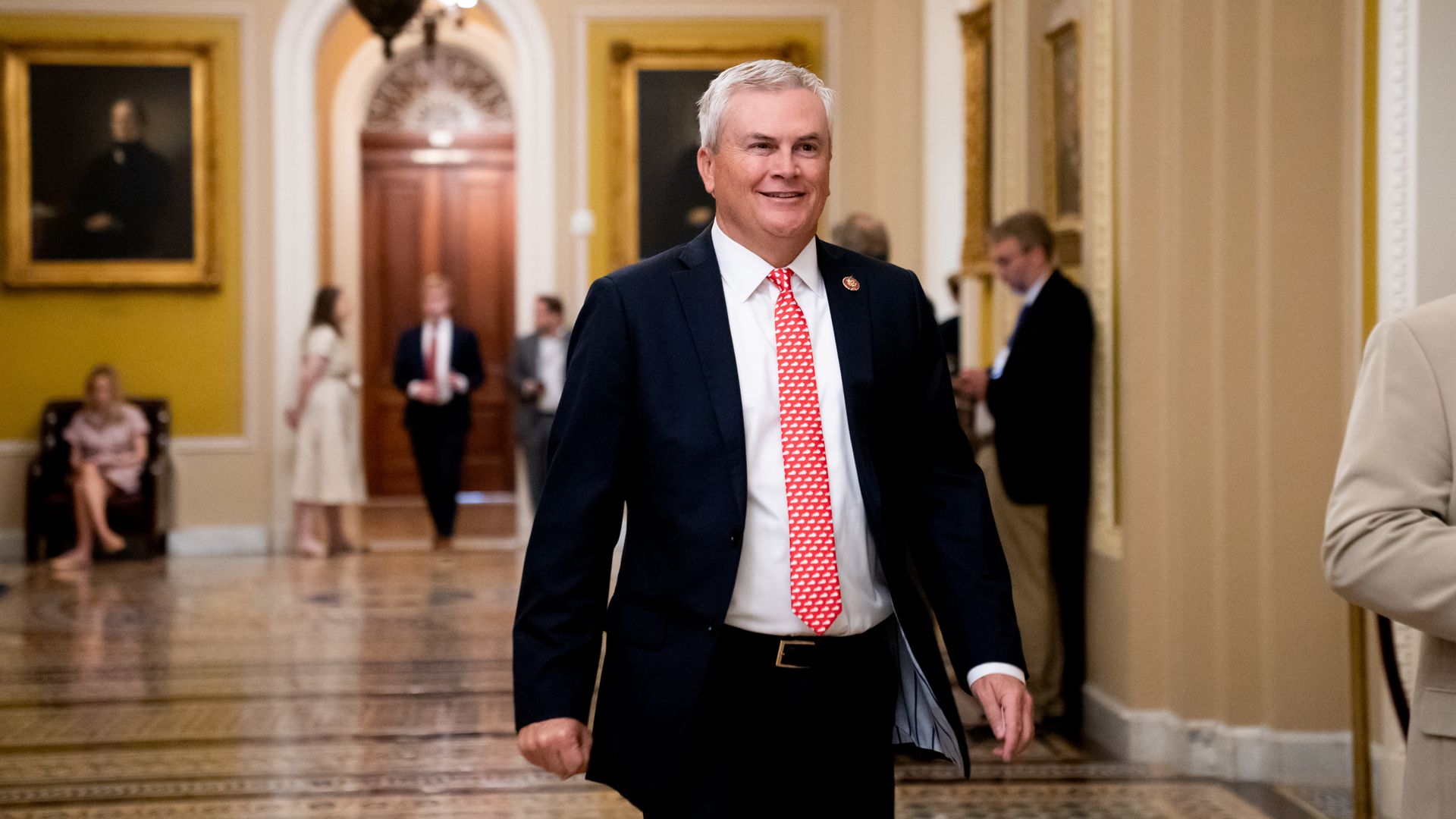 James Comer wearing a black suit and walking through a yellow and white hallway.