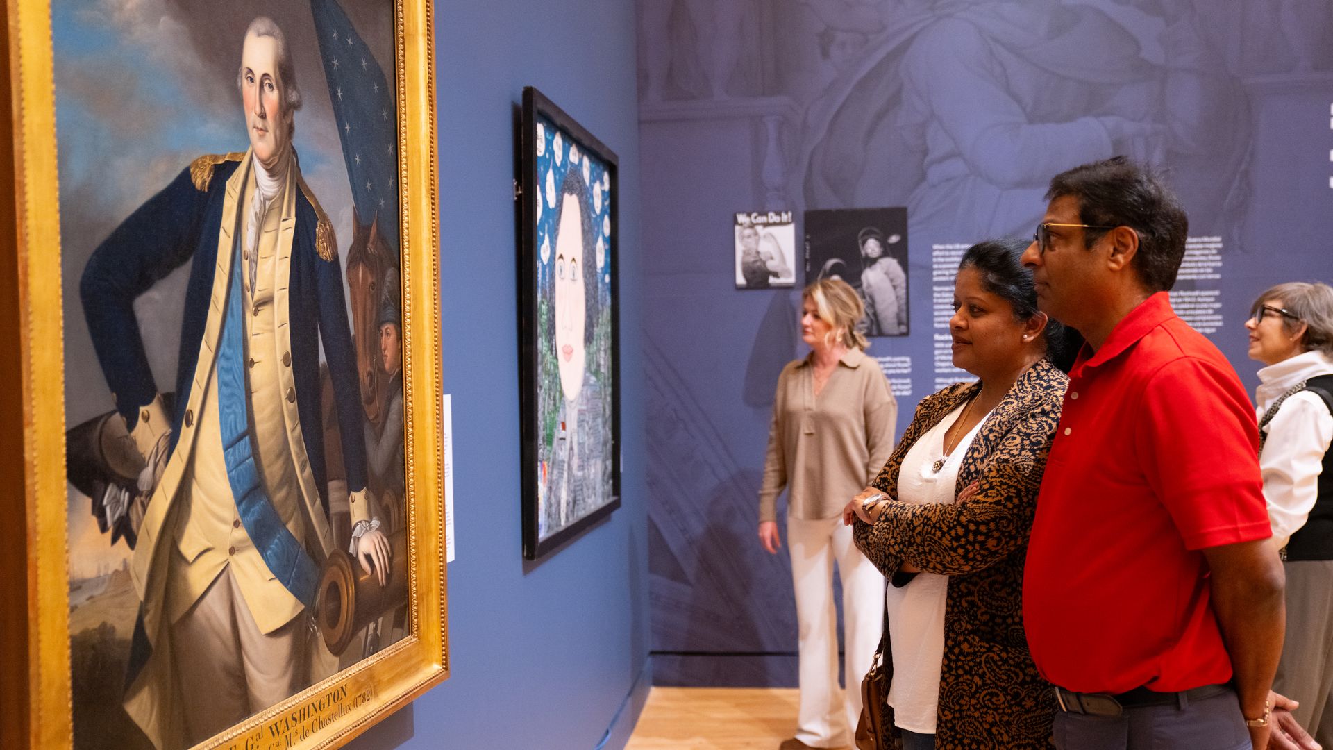 Visitors look at a framed portrait of George Washington displayed on a blue museum gallery wall.