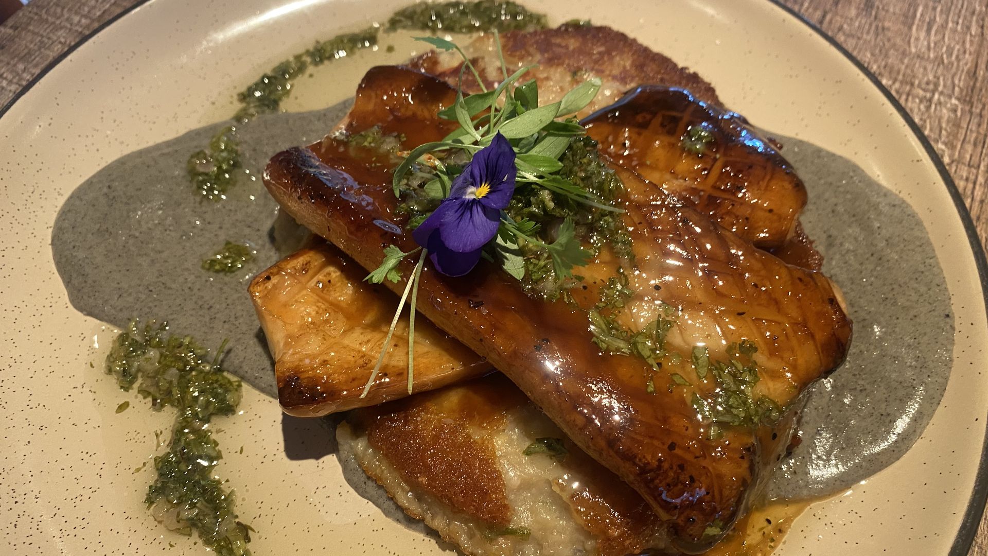 Grilled glazed oyster mushroom on a cream-colored plate with black sauce, green herb garnish, microgreens, and a purple edible flower, on a wooden table.